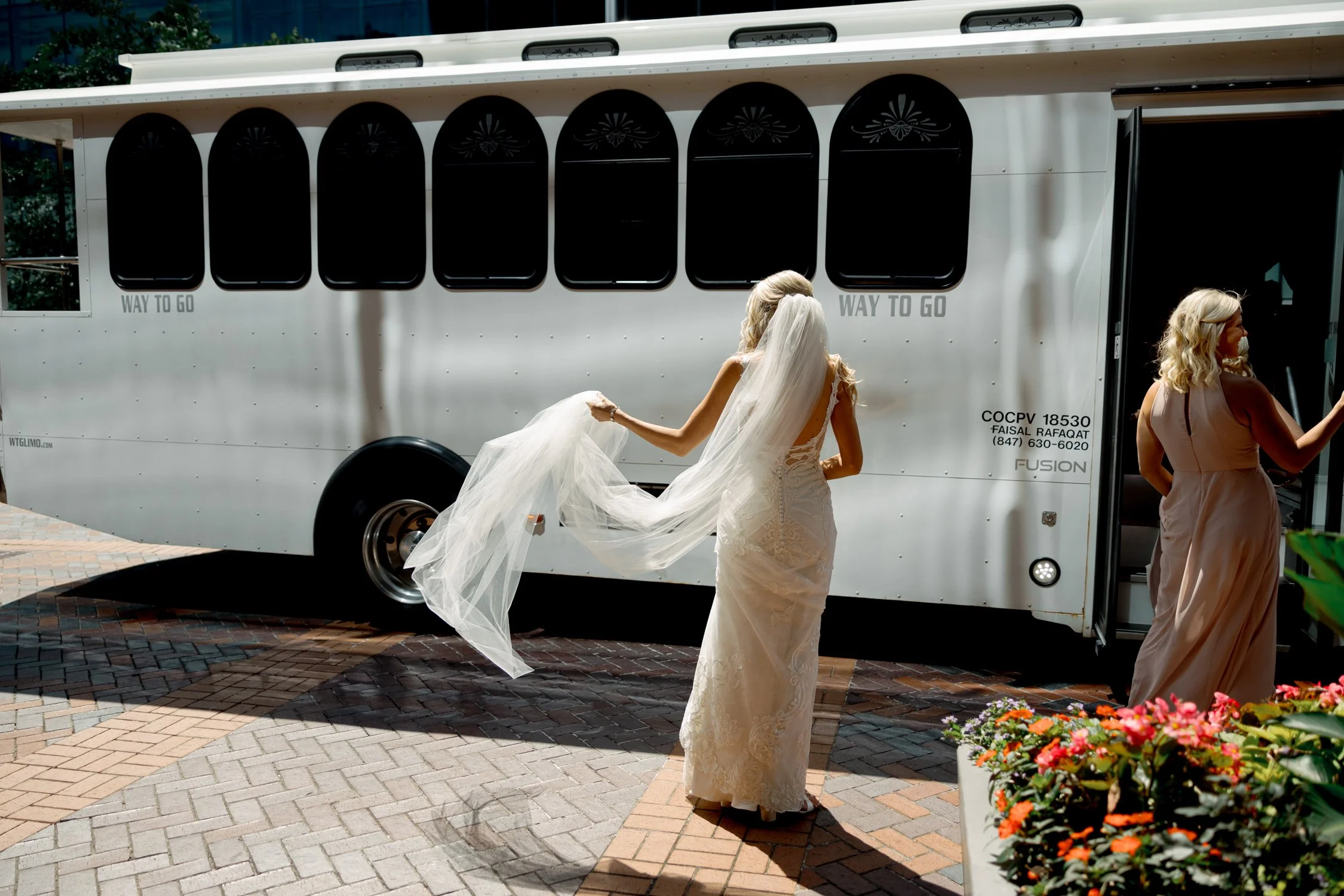 The bride gathers her flowing cathedral veil before stepping onto the wedding trolley, sunlight catching the lace details of her fitted gown outside the venue.