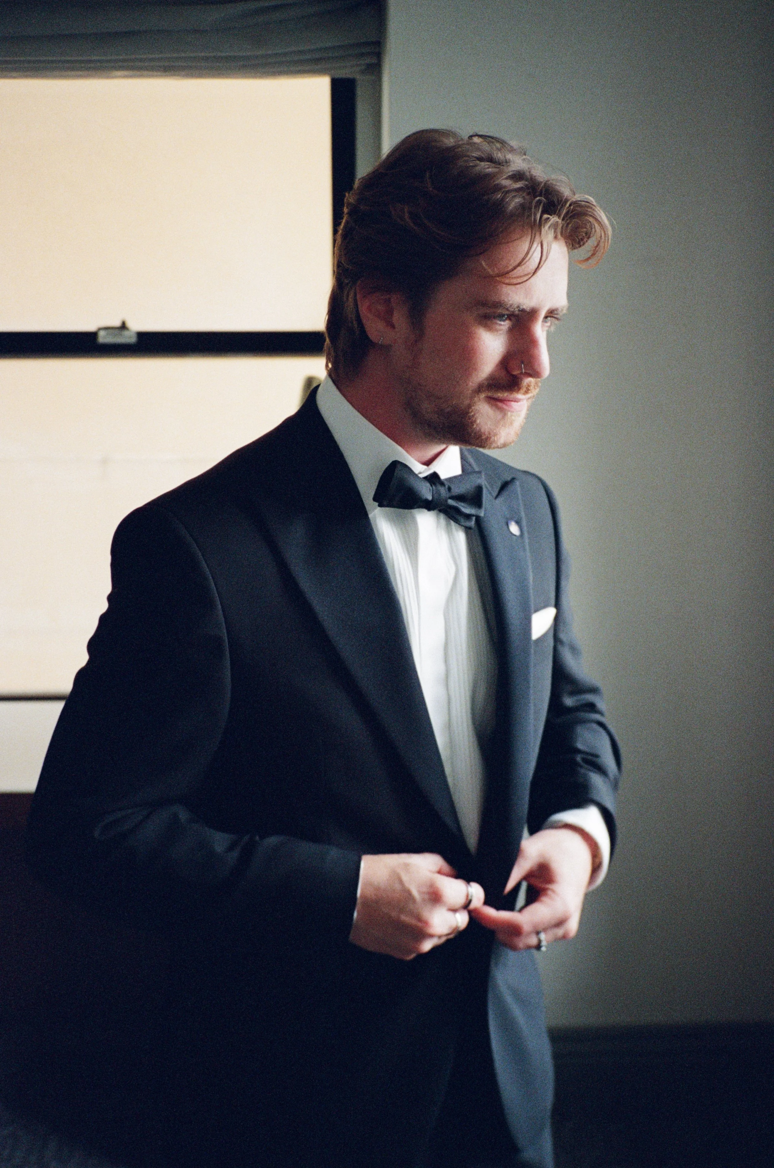 Groom buttons his tuxedo jacket in soft window light while getting ready