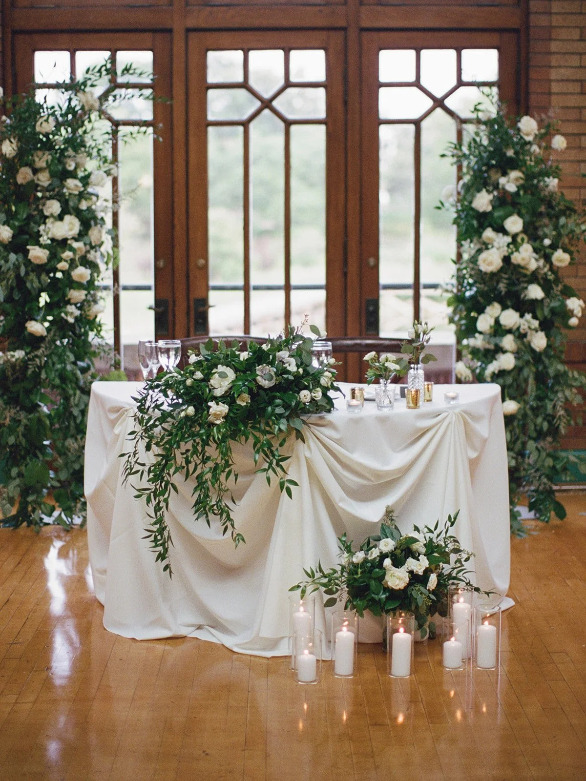 Cafe Brauer sweetheart table — white linen with cascading white rose and greenery garland, pillar candles, and floral arches.