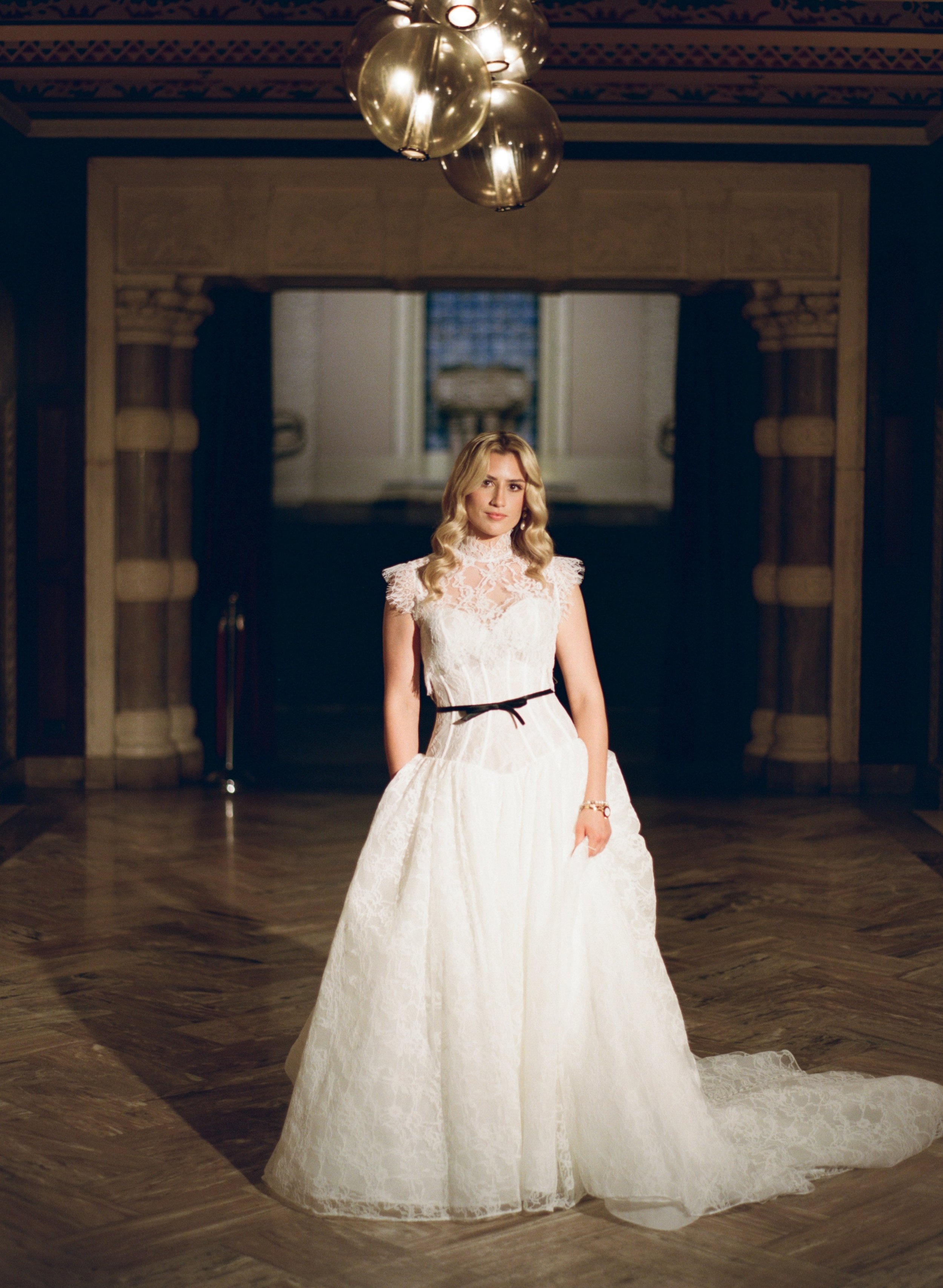Bride in a high-neck lace wedding gown with a black ribbon belt stands in a grand marble hallway beneath a crystal chandelier