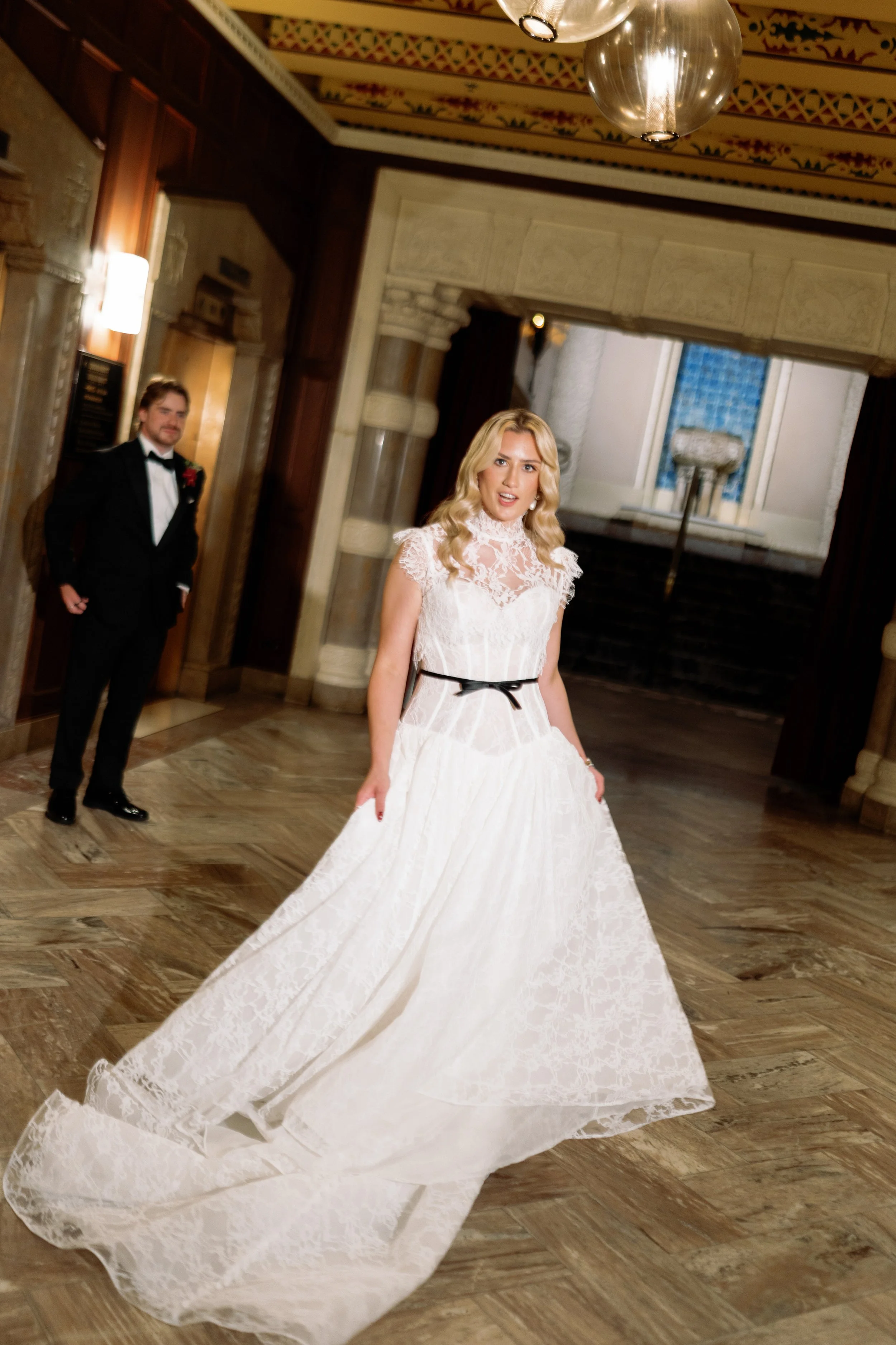 Bride twirls her lace gown train through an ornate hotel hallway as the groom looks on