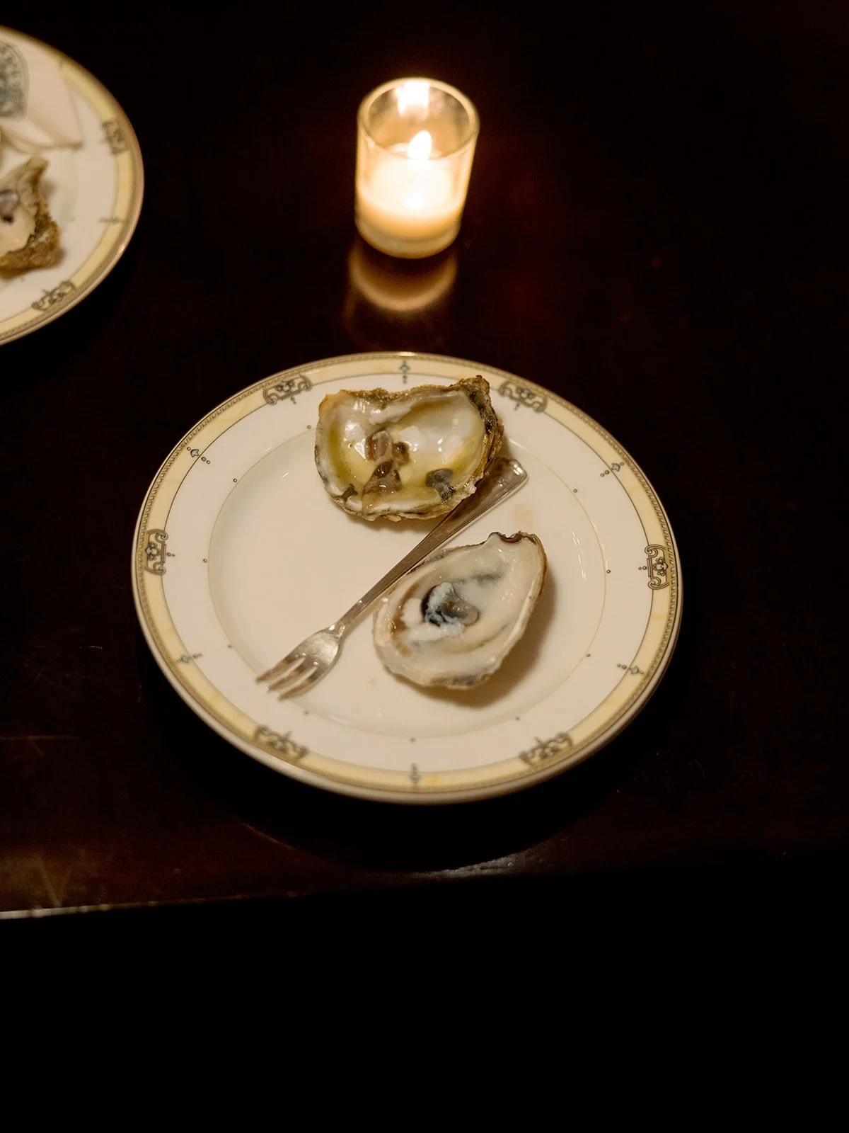 Two fresh oysters on a gold-rimmed University Club china plate, a small cocktail fork laid across them, a votive candle glowing softly in the background. Effortlessly elegant catering detail — shot on film by a Chicago film wedding photographer.