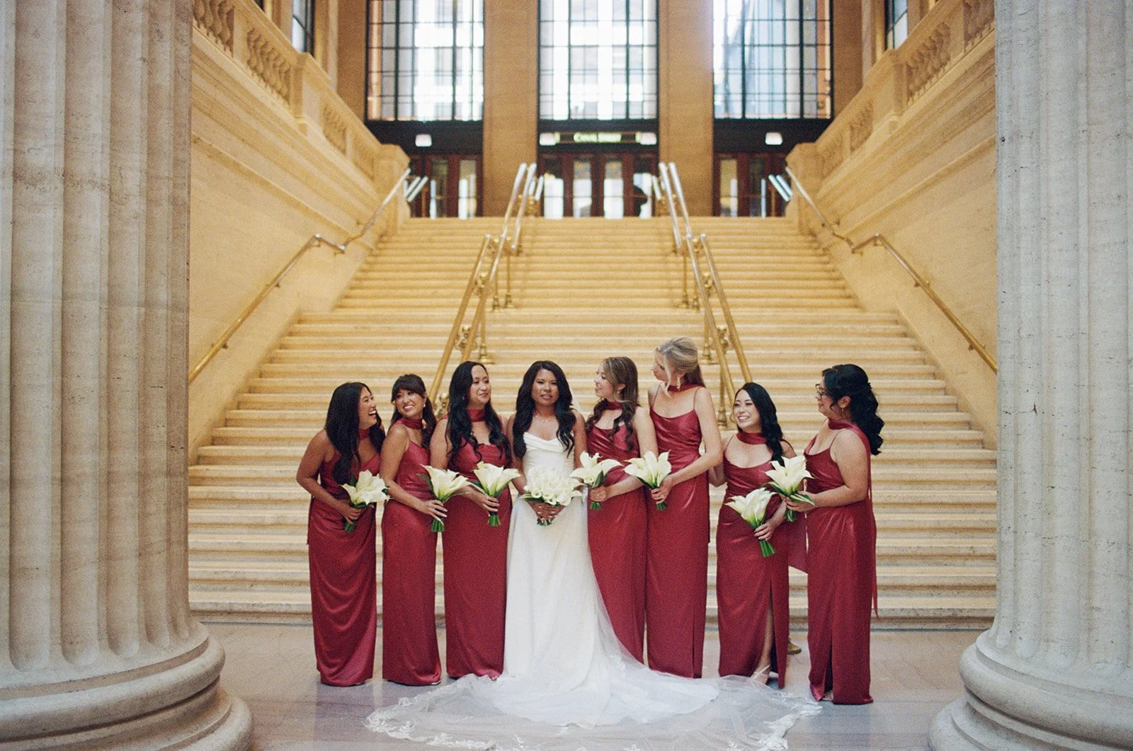 A stunning bridal party portrait on Union Station's grand marble staircase. Bridesmaids in burgundy satin gowns surround the bride in white. Shot on film by a Chicago wedding photographer.