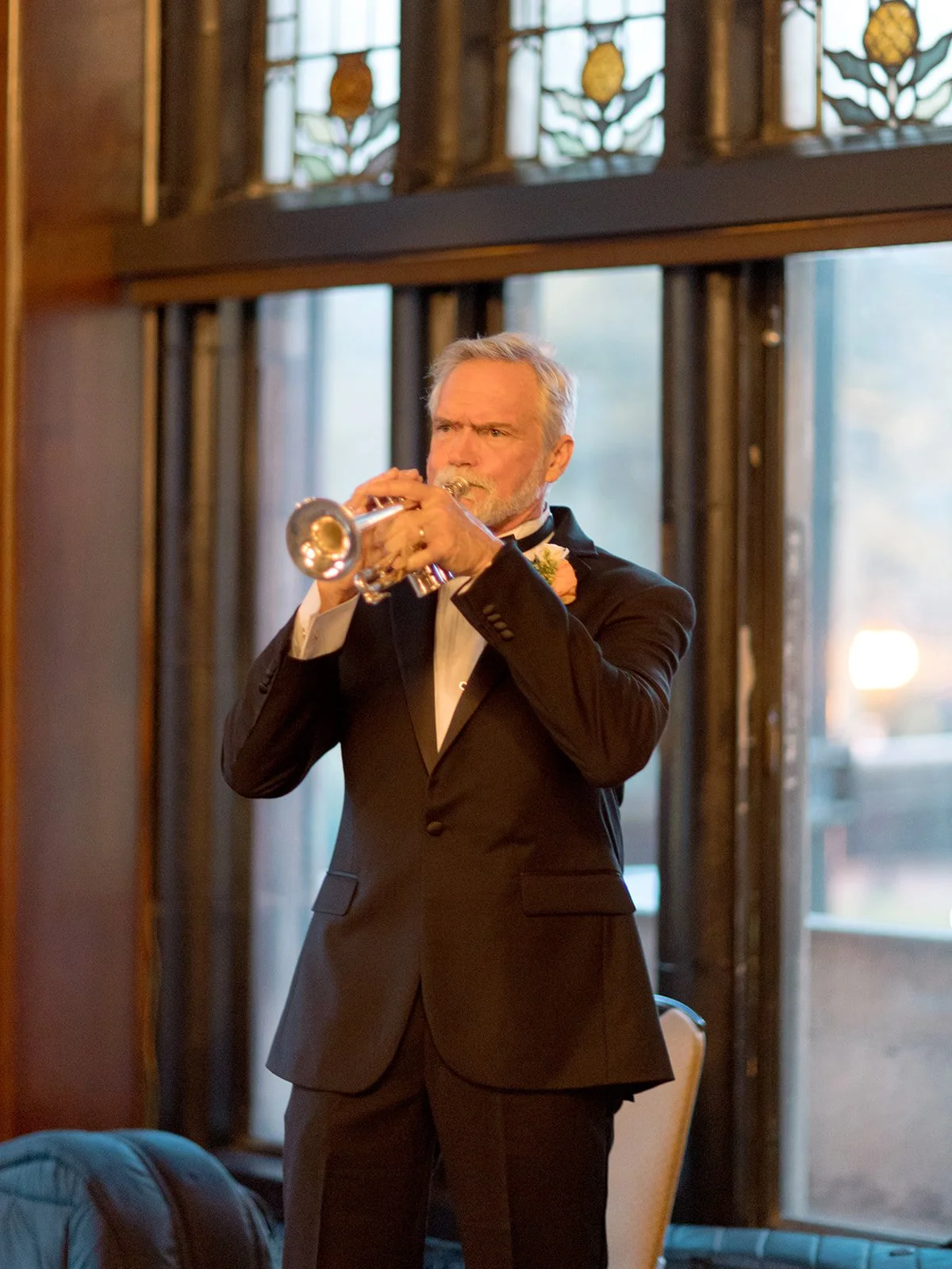 A tuxedoed musician playing a trumpet solo in front of the University Club Chicago's glowing stained glass windows — an unexpected, elegant moment of live entertainment during the reception.