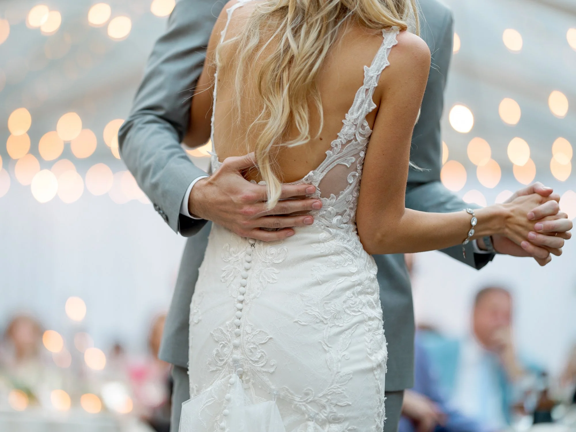 A stunning detail of the bride's lace open-back gown during the first dance, the groom's hands wrapped gently around her as string lights glow above.