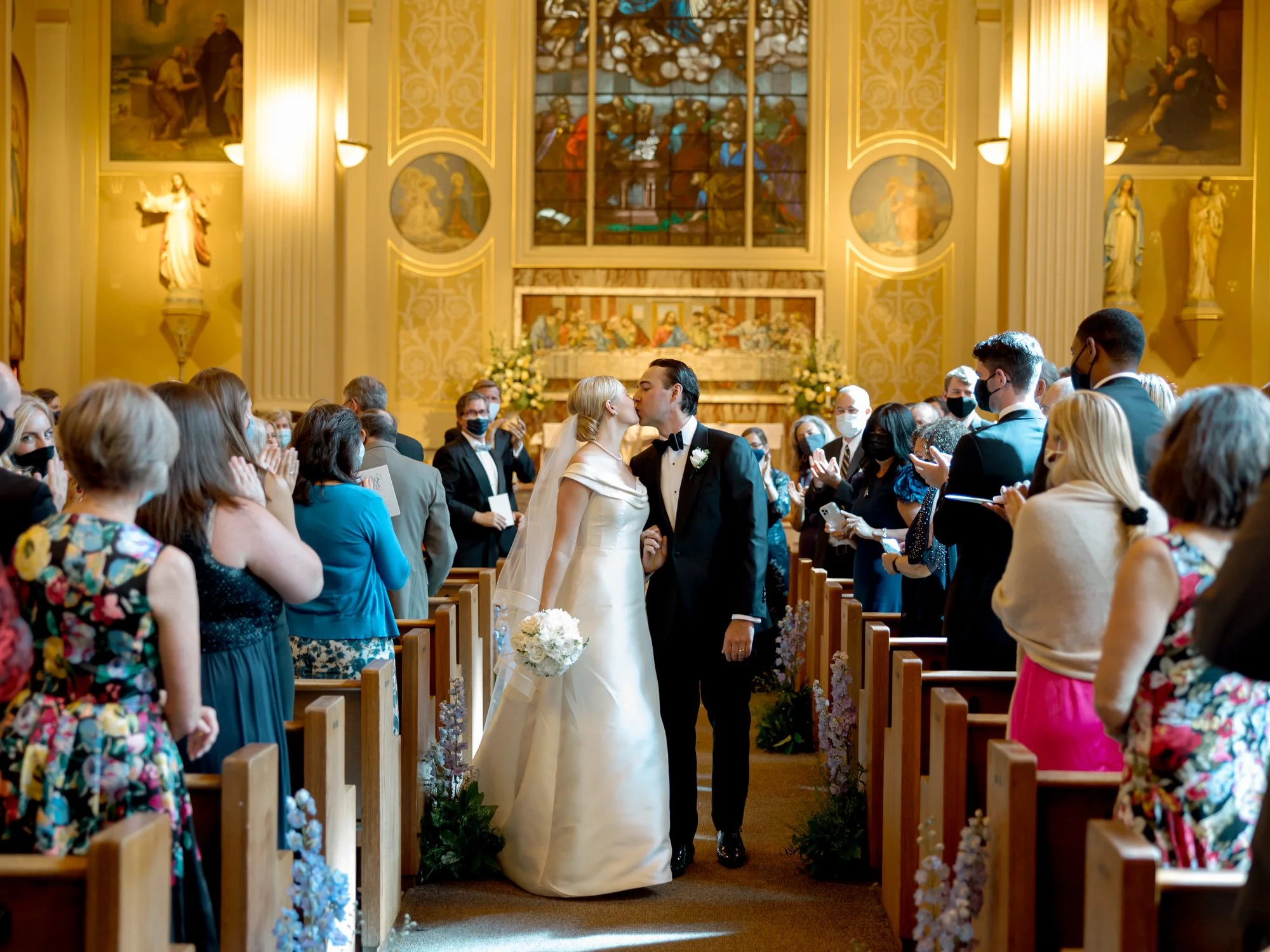 Bride and groom pause to kiss mid-recessional, stained glass windows glowing at the far end of the chapel, guests clapping on every side around them.
