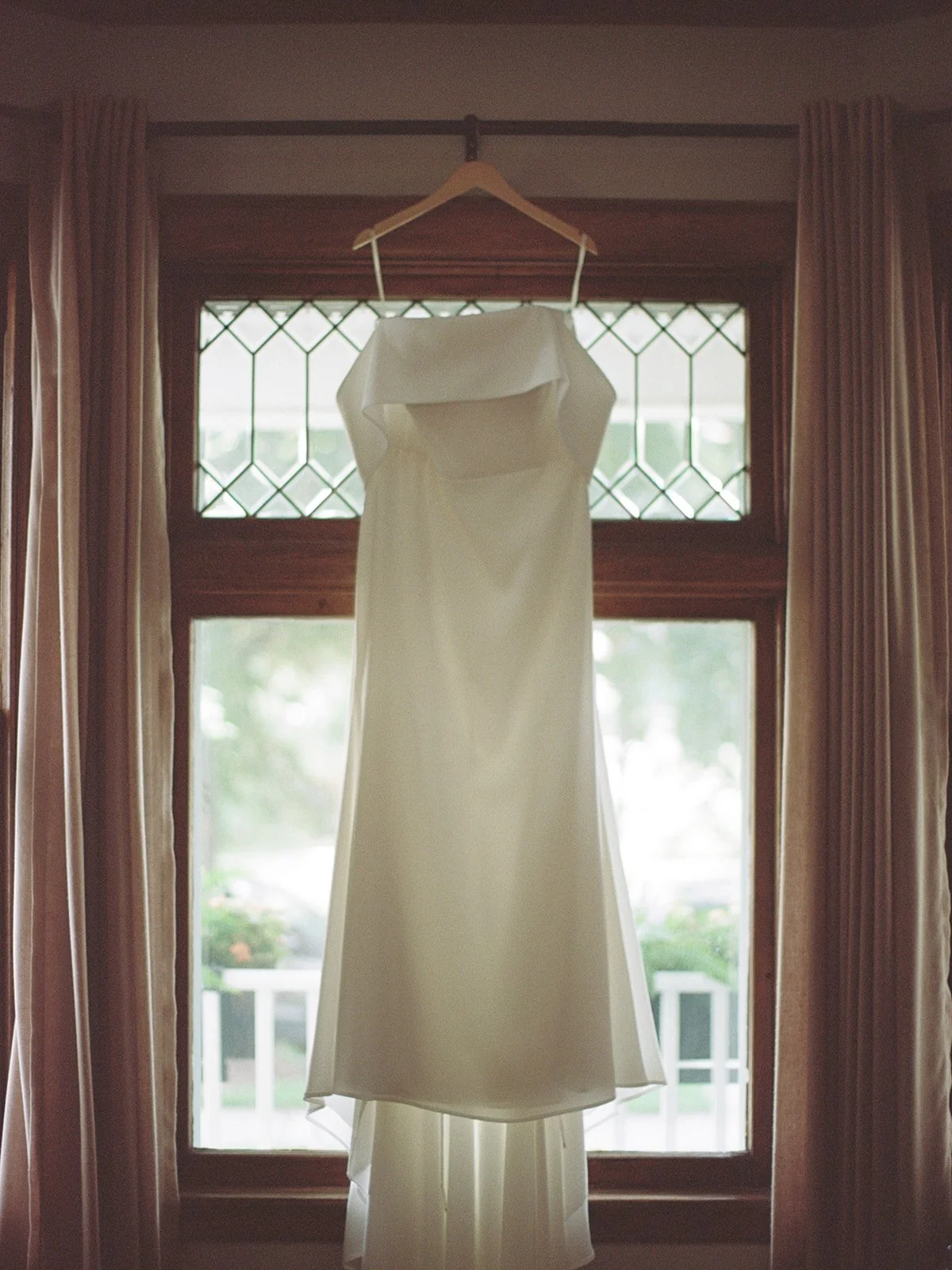 Ivory off-the-shoulder wedding gown hanging in a sunlit window with leaded glass panes during bridal getting ready.