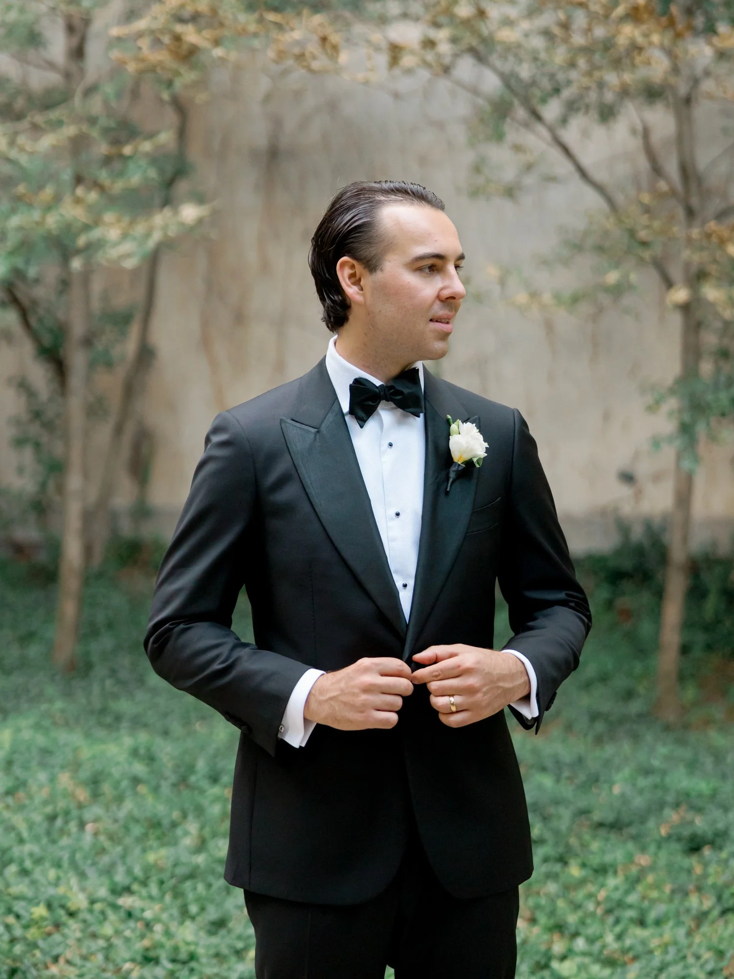 A classic groom portrait: sharp black peak-lapel tuxedo, a white boutonniere, and a lush ivy-covered garden wall behind him. Shot on film in Chicago.