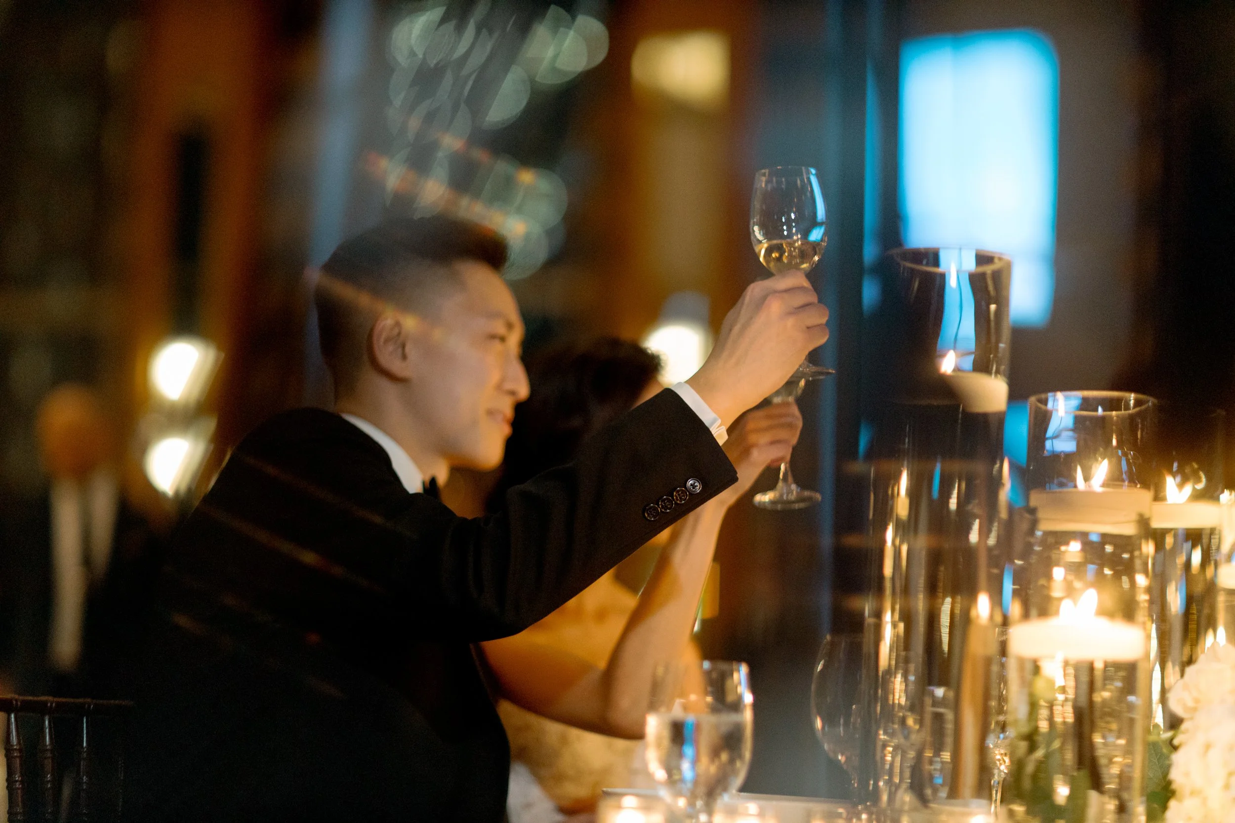 Groom holding up a glass of wine at a candlelit reception table at The Peninsula Chicago, warm evening light