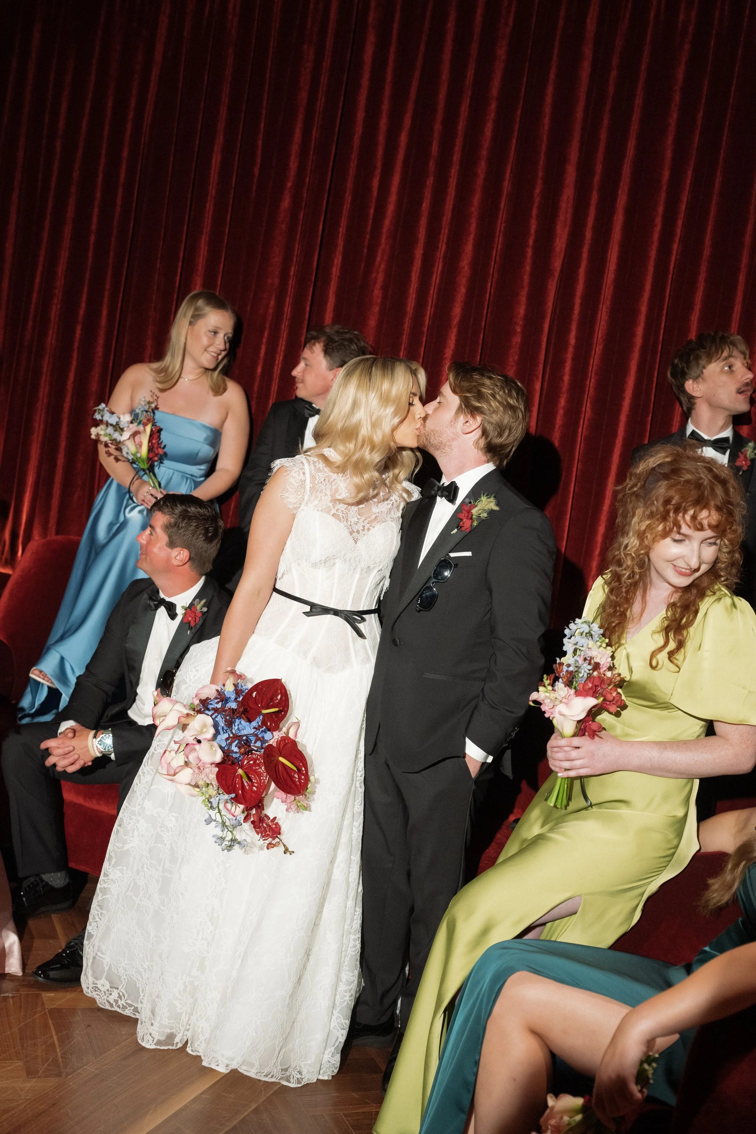 Bride and groom kiss while surrounded by their bridal party on the stage inside The Wellsley's red velvet lounge