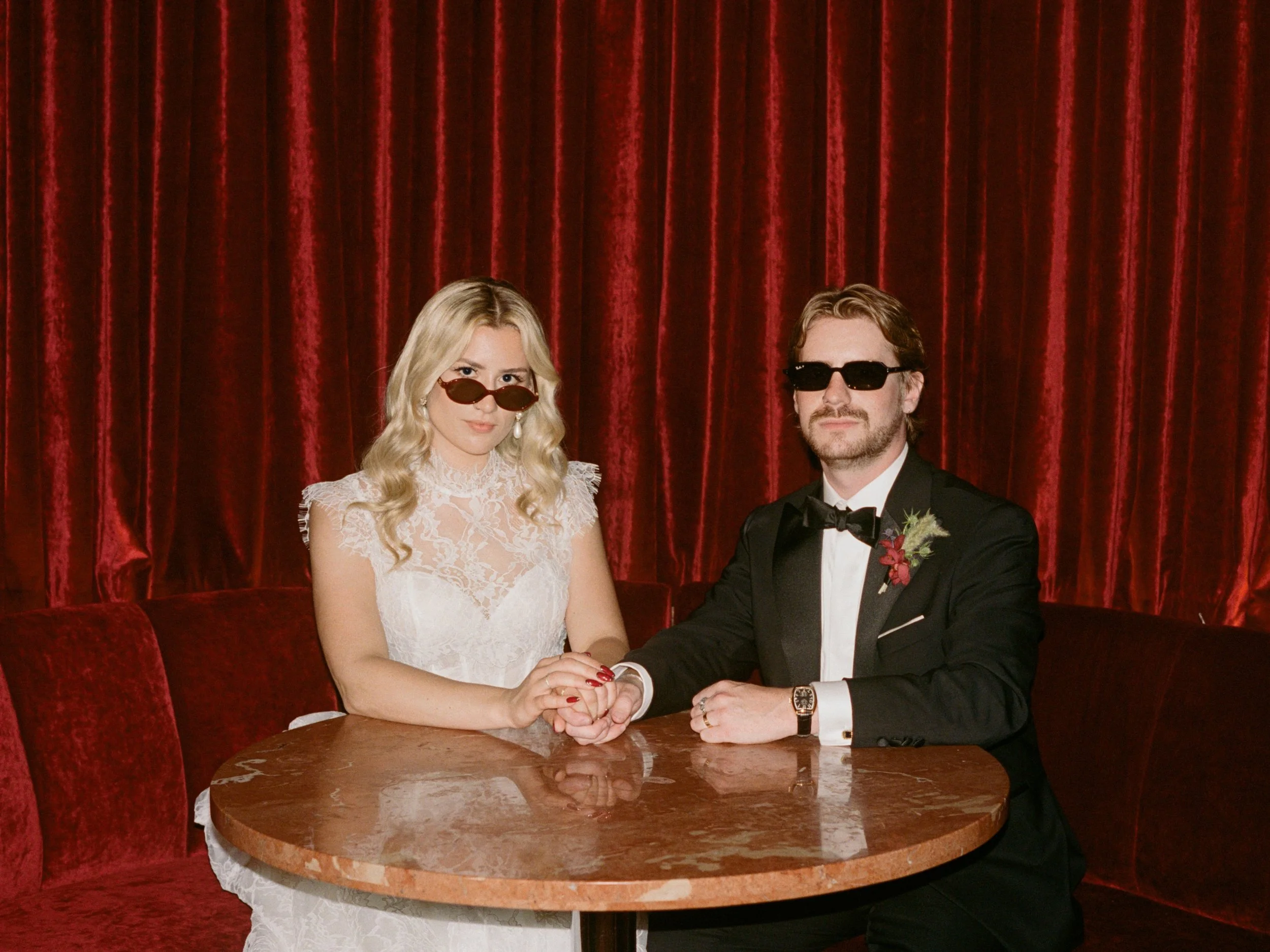 Bride and groom pose together in sunglasses at their round table inside The Wellsley's red velvet lounge