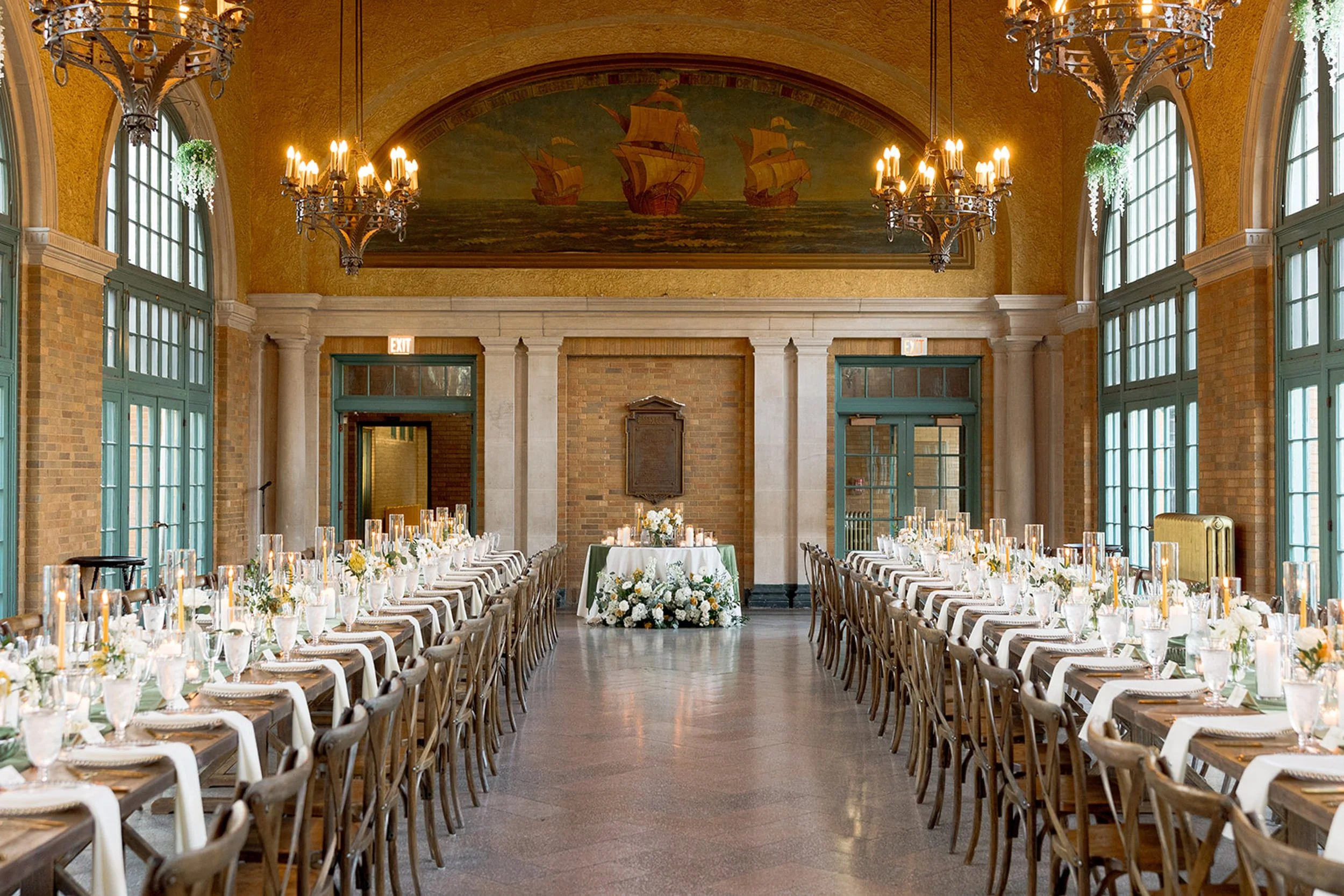 The full grand interior of Columbus Park Refectory in all its glory — a vintage nautical ship mural overhead, twin rows of chandeliers, long farm tables set in perfect symmetry for the wedding reception