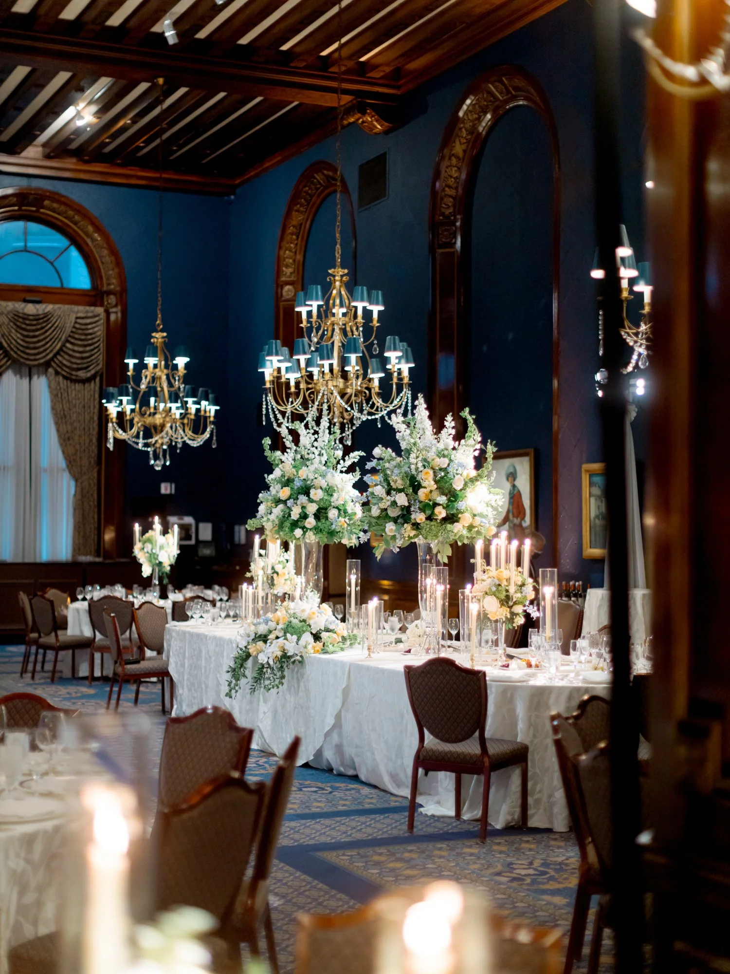 The Union League Club reception room fully set with deep navy walls, ornate gold chandeliers, and towering white floral arrangements at the head table.
