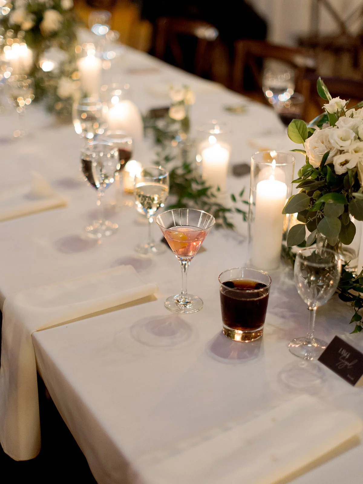 Close detail of a reception table at Cafe Brauer — cocktails, pillar candles, and white florals glowing in candlelight.