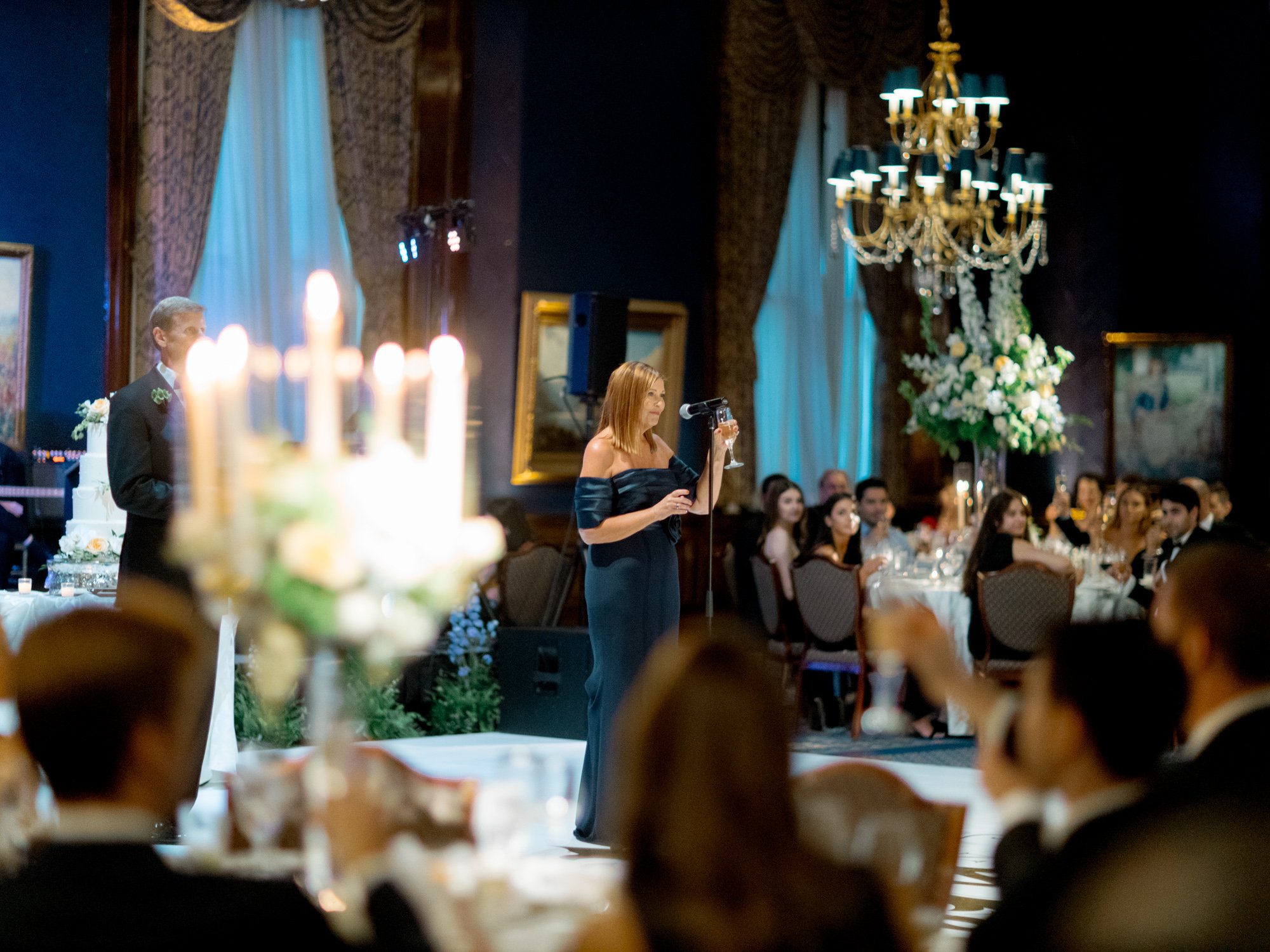 A woman in a chic navy off-shoulder gown delivers her toast at the microphone, the full candlelit Union League Club grand ballroom listening behind her.
