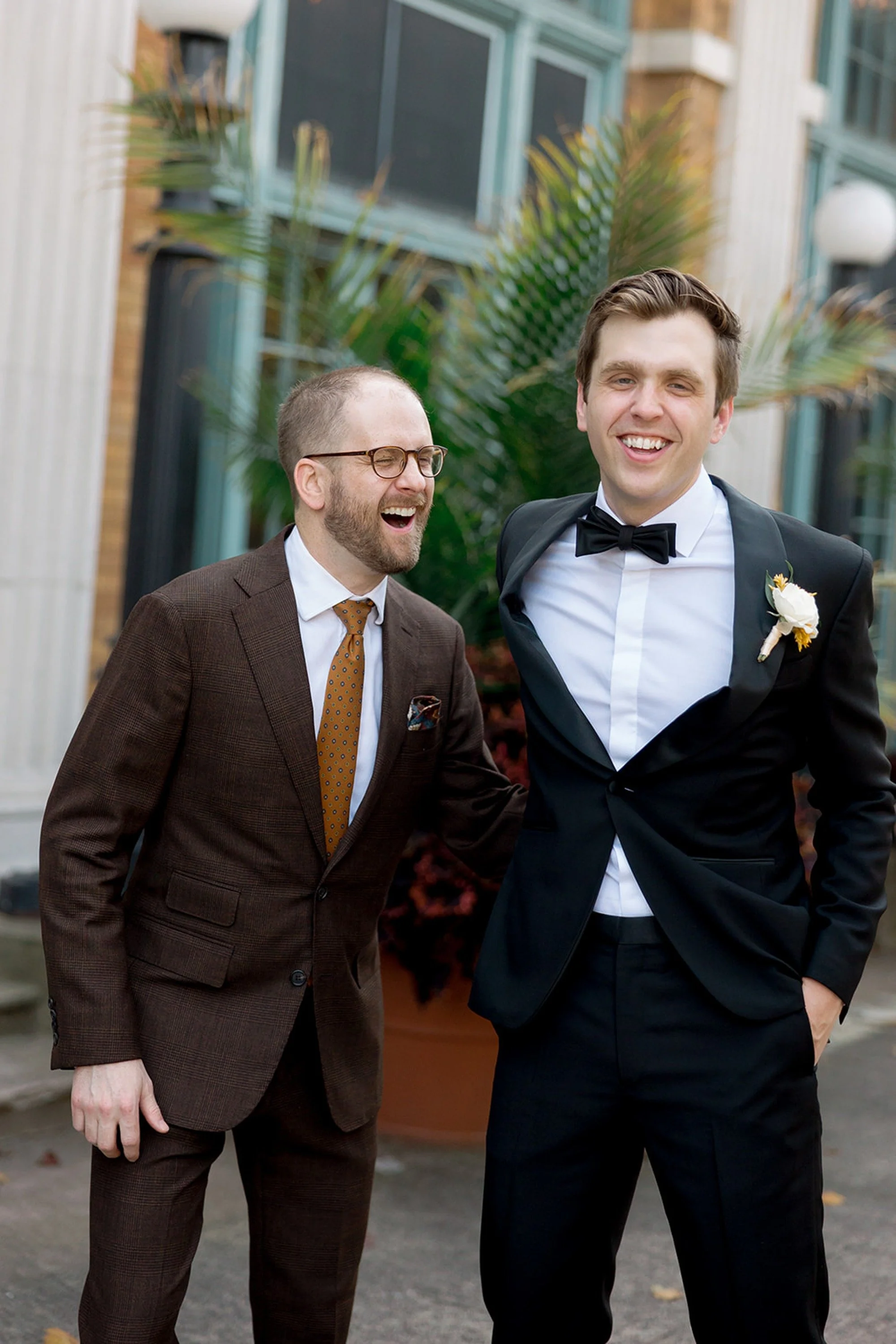The groom and his groomsman share a genuine, unguarded laugh outside Columbus Park Refectory — a candid portrait full of camaraderie on the wedding day