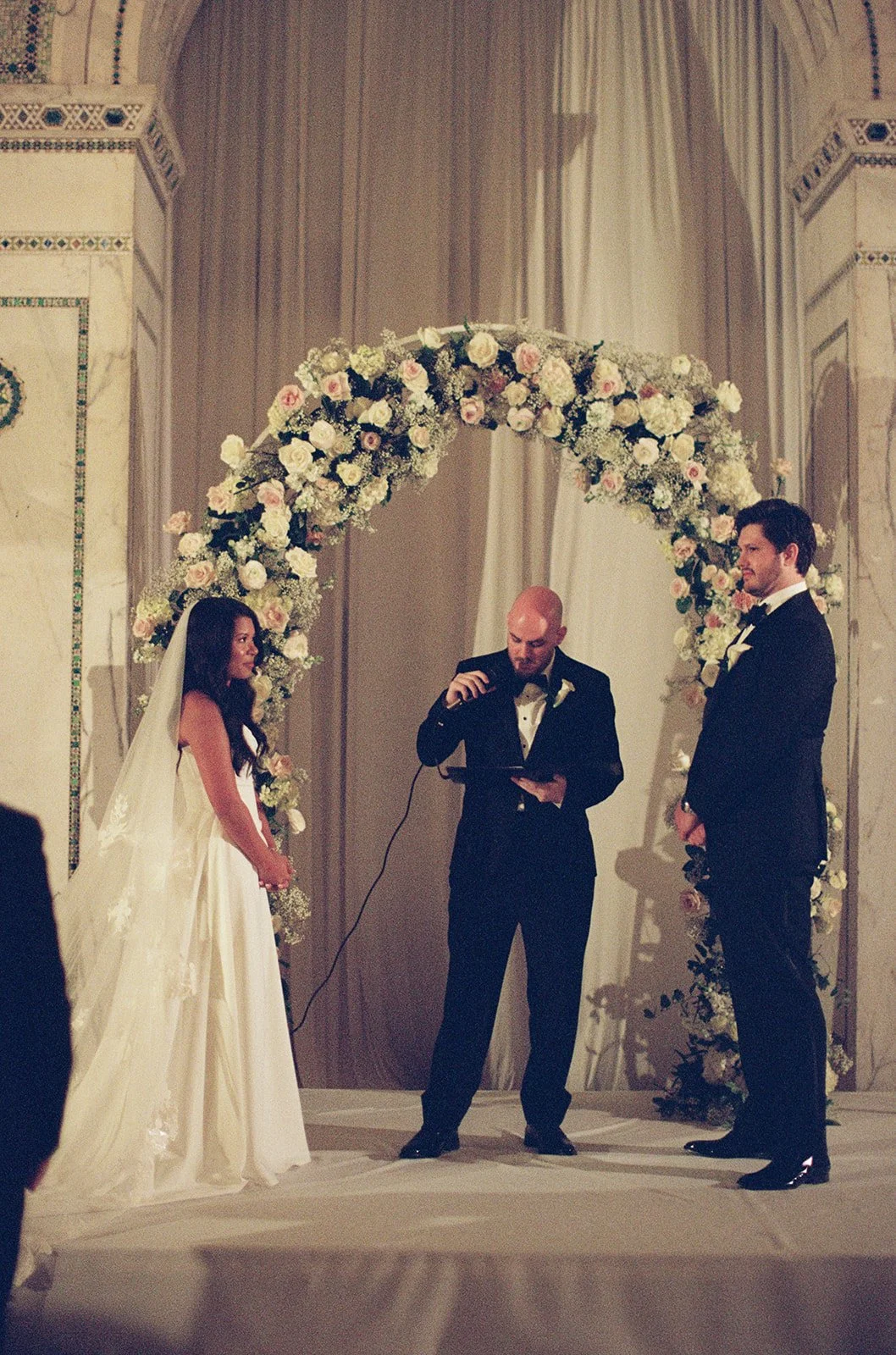 Sarah and Brennan exchange vows under a stunning white floral arch during their ceremony at Chicago Cultural Center. The bride in a sleek white gown and groom in black tuxedo share this intimate moment. Shot on film by a Chicago wedding photographer.