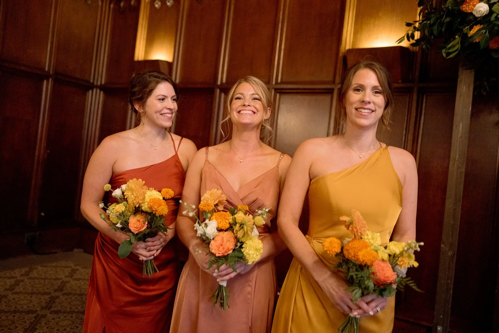 Three bridesmaids in rust, camel, and mustard gowns sharing a genuine laugh in the University Club Chicago's wood-paneled hallway, bouquets in hand. The autumn color palette feels completely at home in this historic space