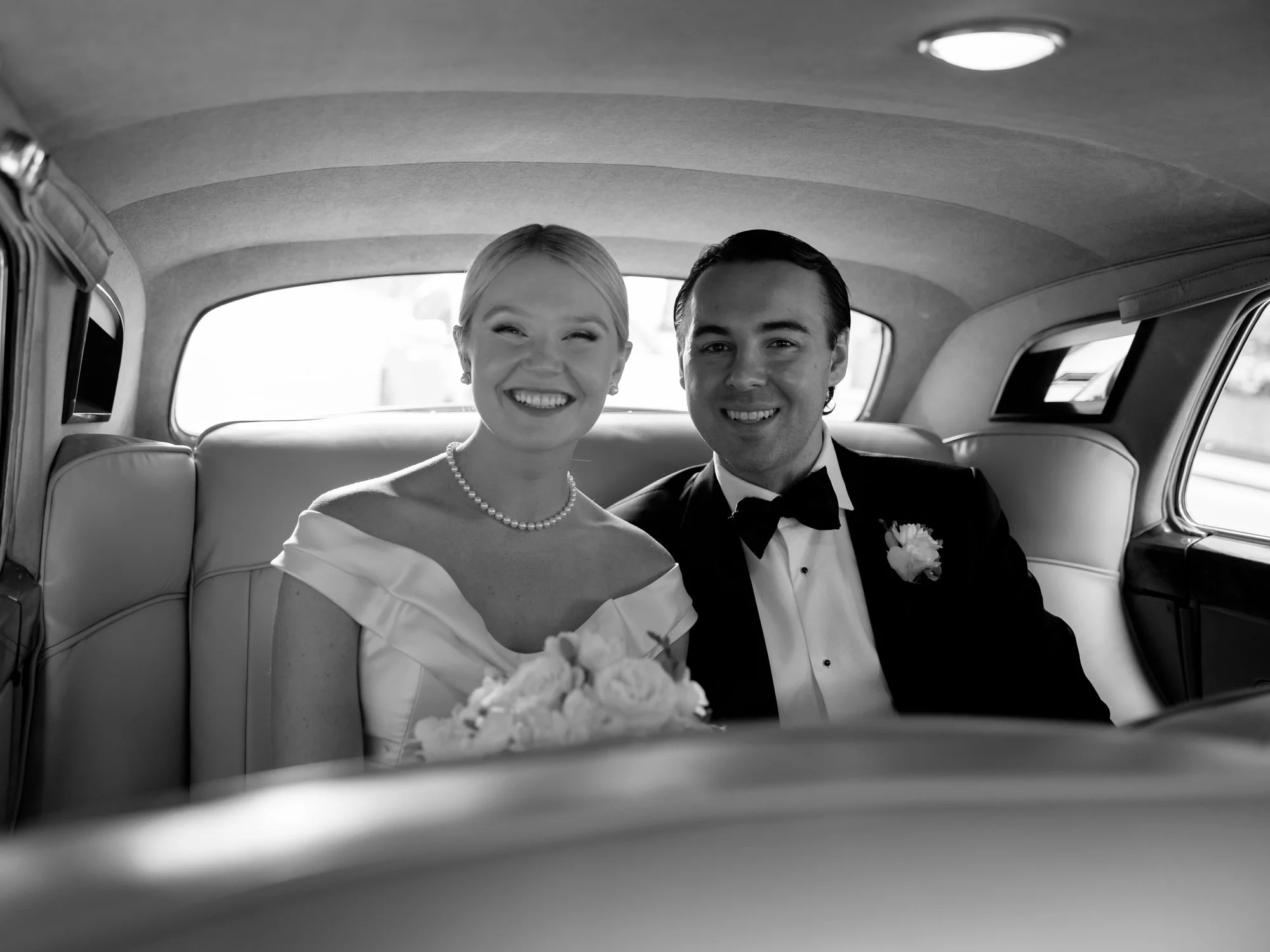 Bride and groom beam side by side in the backseat of a vintage Rolls-Royce in black and white — just married, the whole evening still ahead of them.