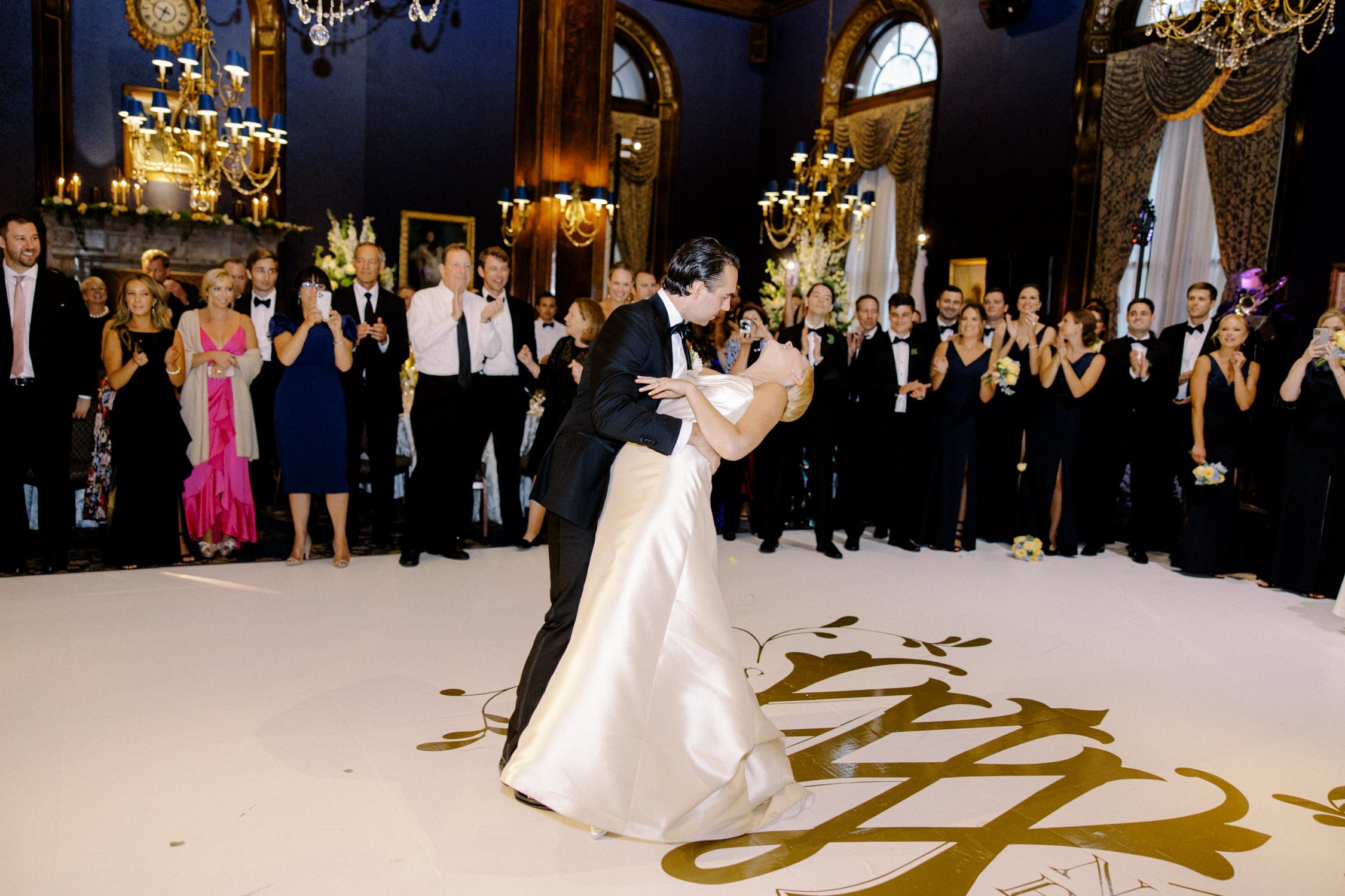 The groom dips the bride at the close of their first dance, the custom gold monogram floor beneath them and guests cheering in a full circle around them.
