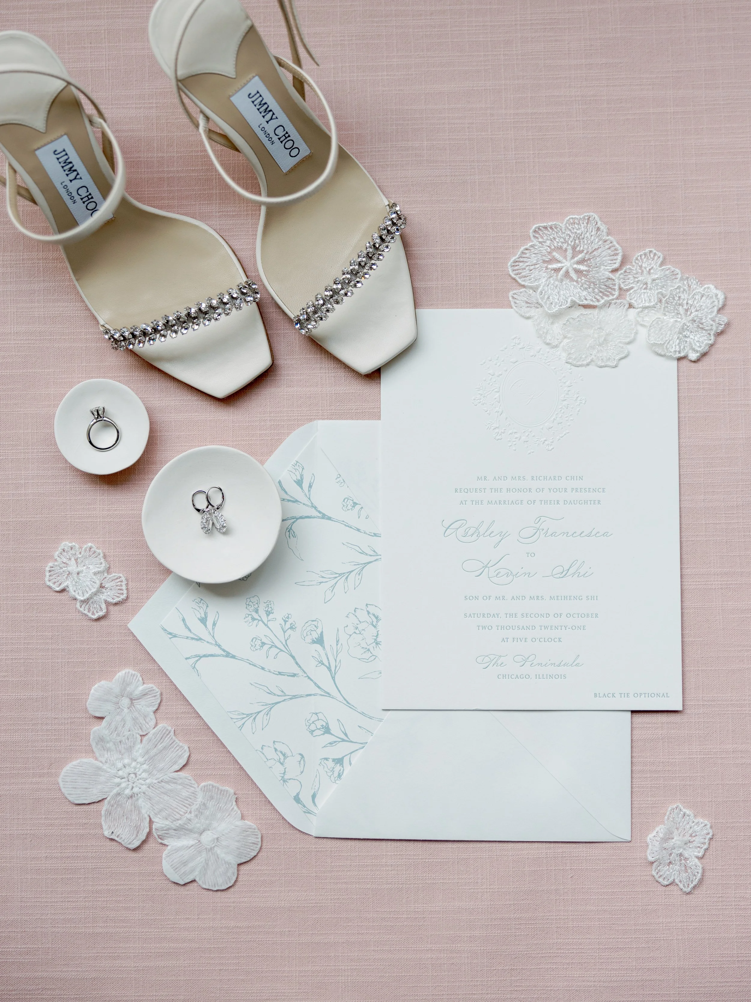 Flat lay of Jimmy Choo heels, Ashley and Kevin letterpress invitation, rings and lace floral details on blush linen