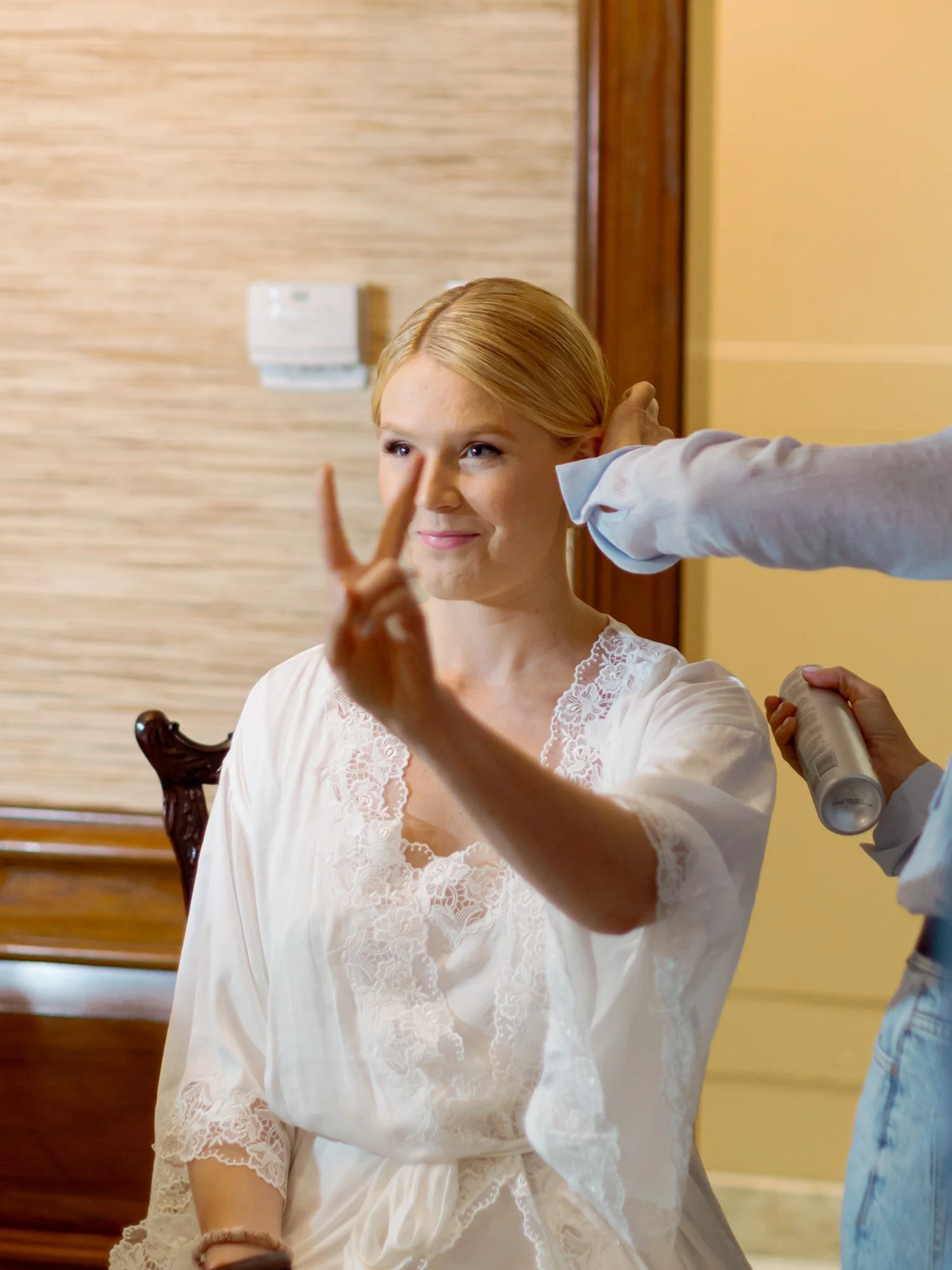 Bride in her white lace robe flashes a peace sign mid-hair appointment at the Union League Club, completely at ease and entirely herself on her wedding day.
