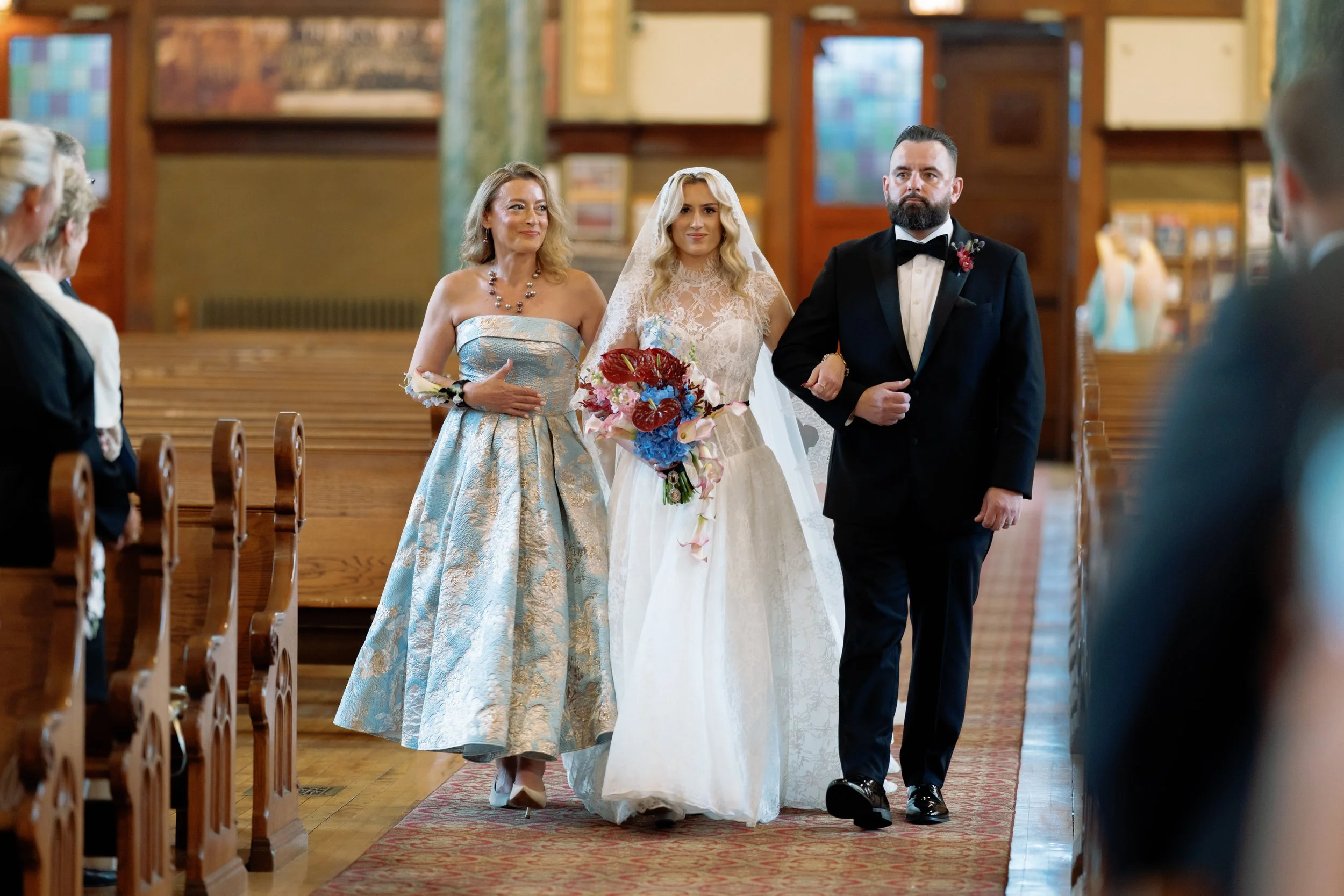 Bride walks down the aisle escorted by her mother, holding a vivid red and blue bouquet