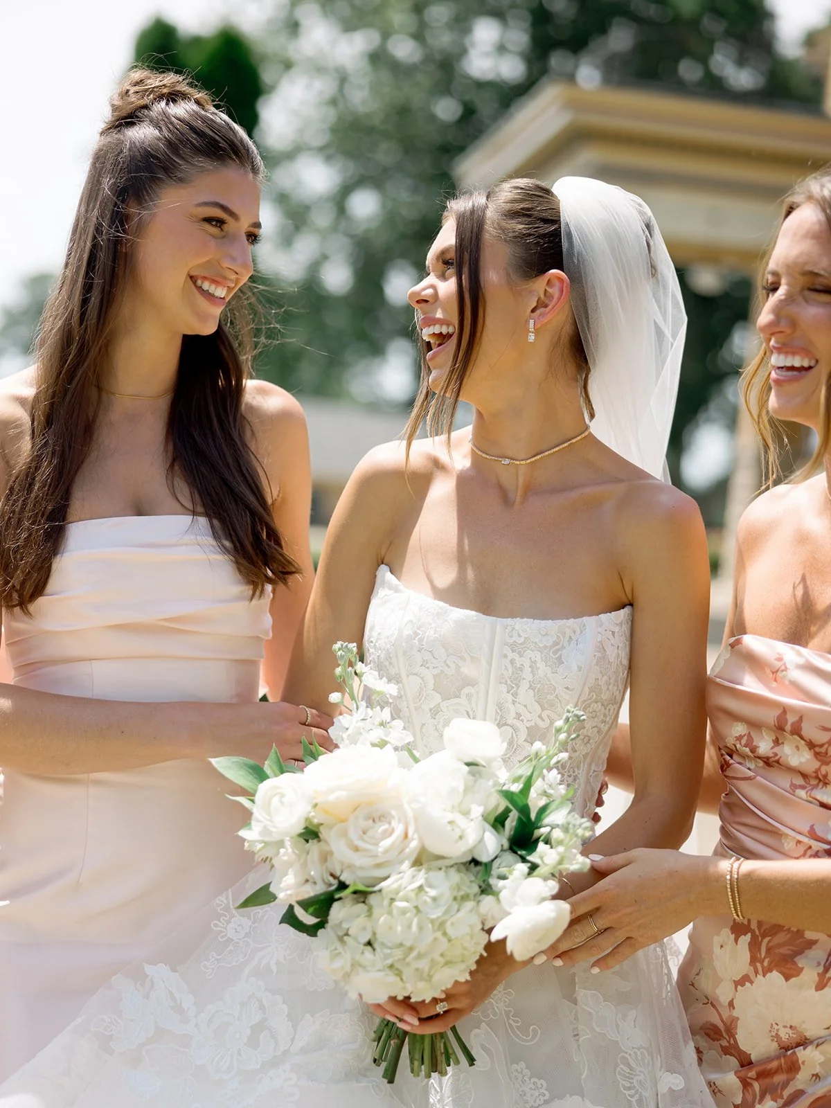 Candid film portrait of a laughing bride in a lace strapless gown with bridesmaids in blush dresses, holding a white rose bouquet outdoors, photographed by Chicago film wedding photographer Louie Abellera.