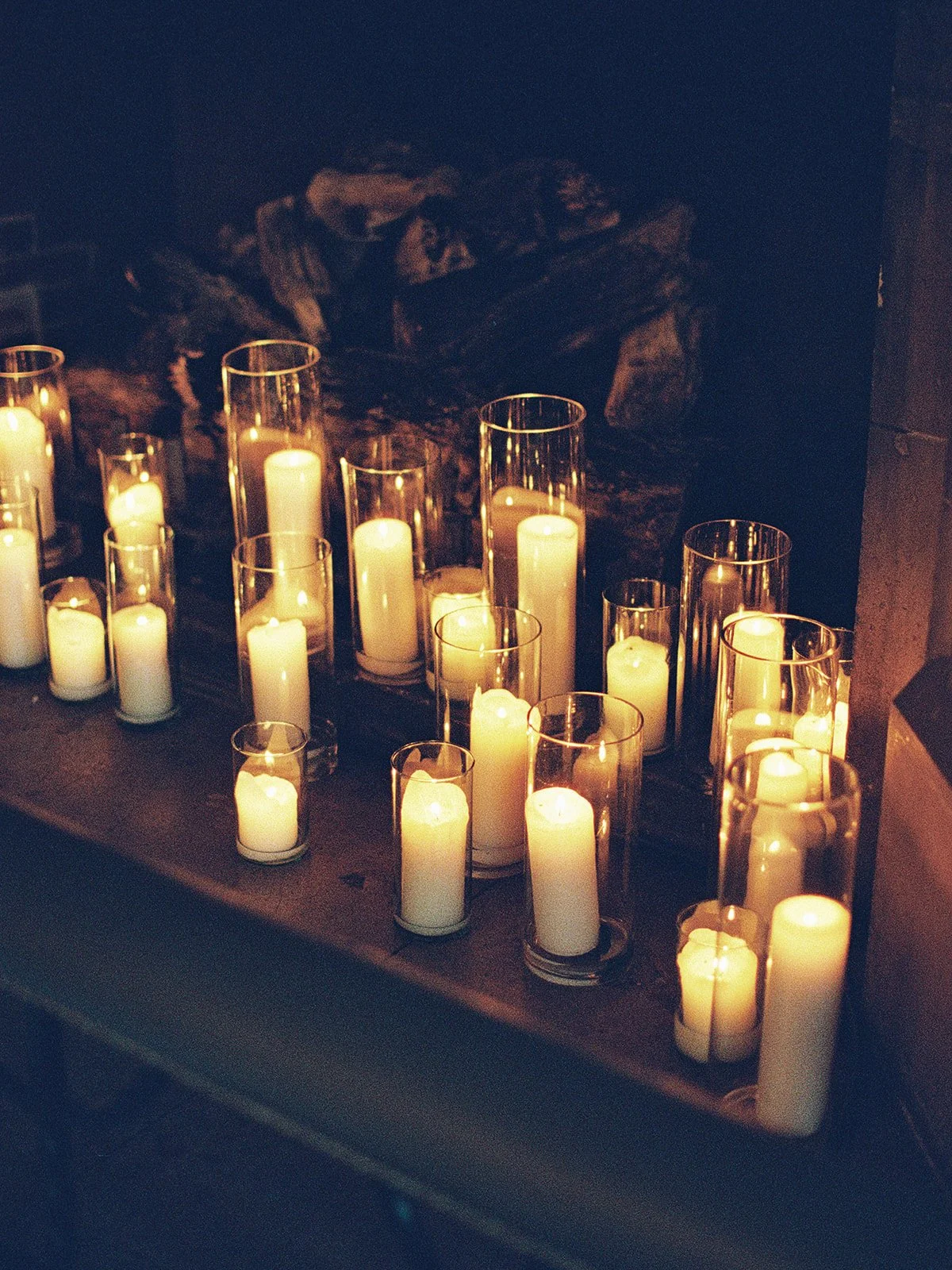 Rows of pillar candles in glass cylinders arranged across the ceremony fireplace mantle, filling the room with a warm amber glow. A moody, romantic detail from the University Club Chicago ceremony