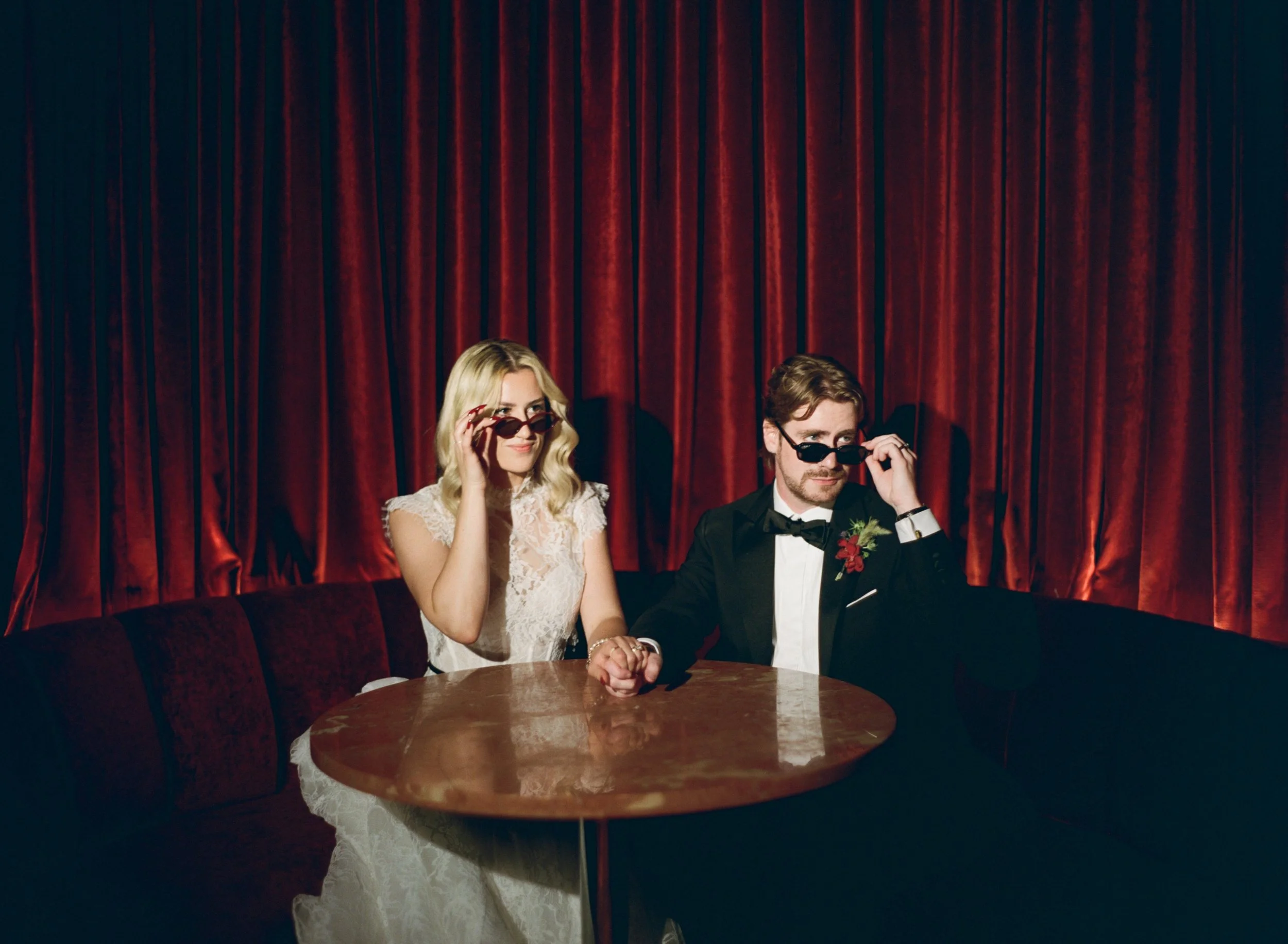 Bride adjusts her sunglasses while holding hands with the groom at their round table in The Wellsley's red velvet lounge