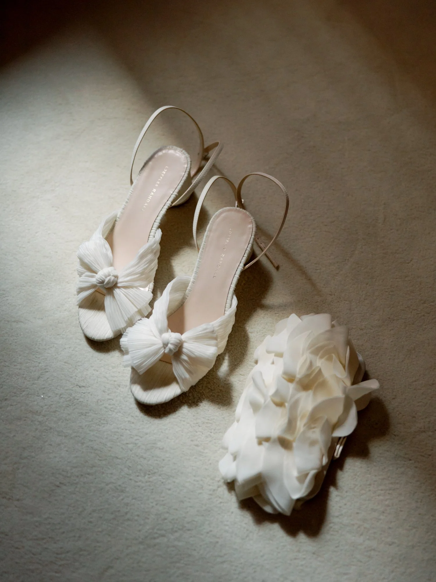 An editorial detail of the bride’s shoes: delicate white silk bow heels beside a sculptural ruffled floral sandal — two considered options, shot on film.