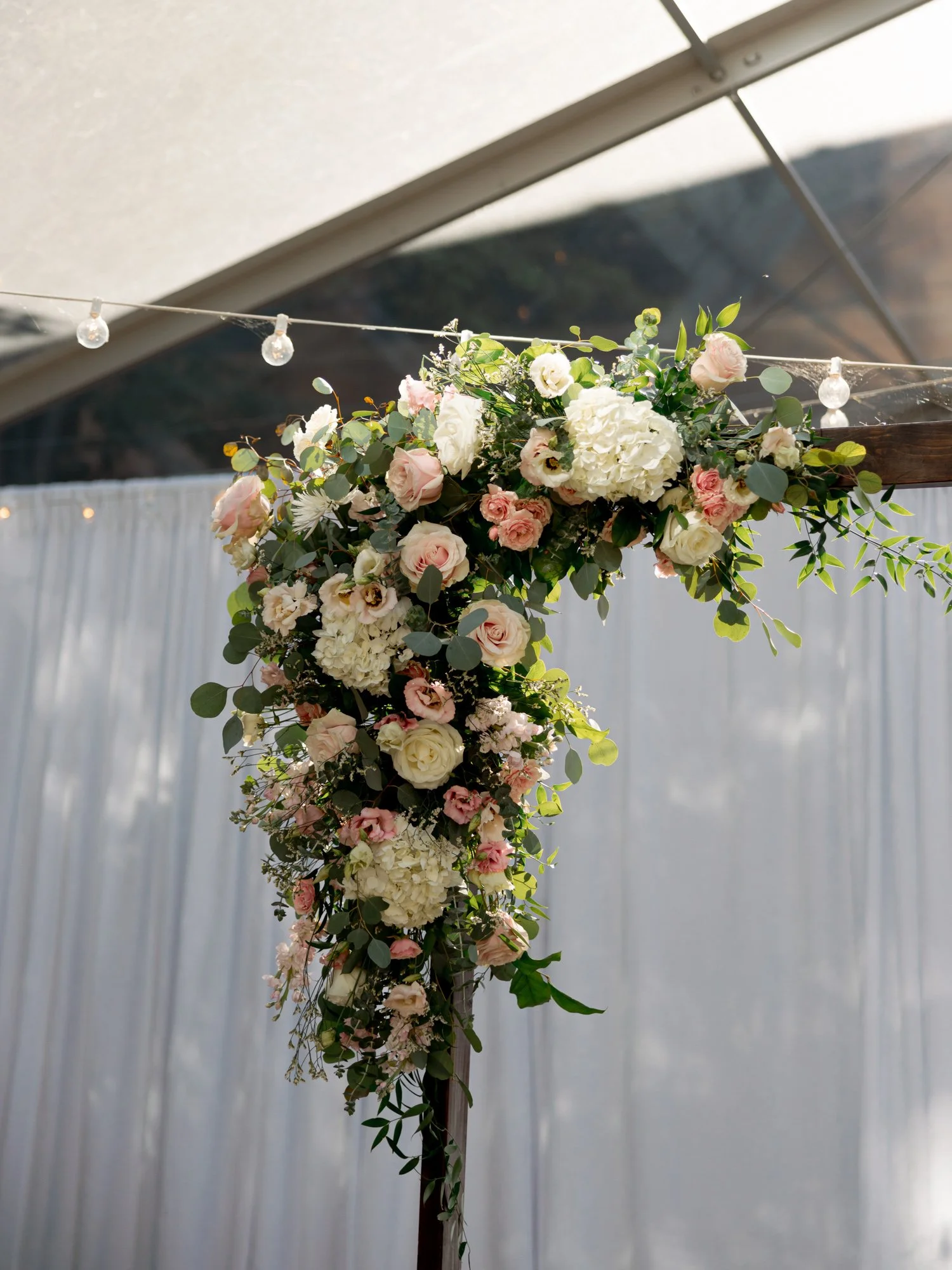 A lush ceremony arch draped in blush roses, white hydrangeas, and trailing eucalyptus at the Chicago Illuminating Company glows under the glass tent.