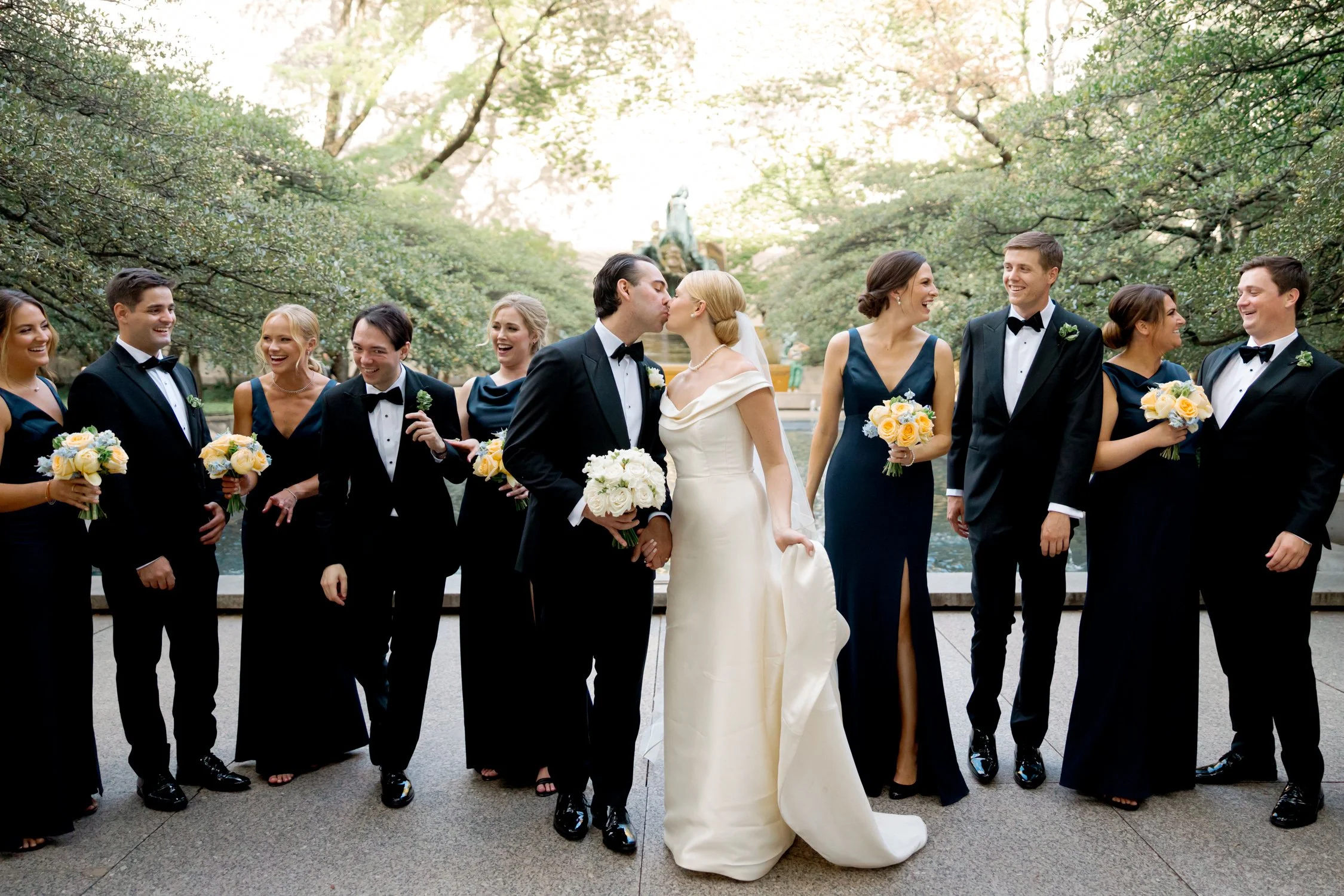 The full wedding party gathers around the kissing bride and groom before a stone garden fountain, captured on film by a Chicago film wedding photographer.
