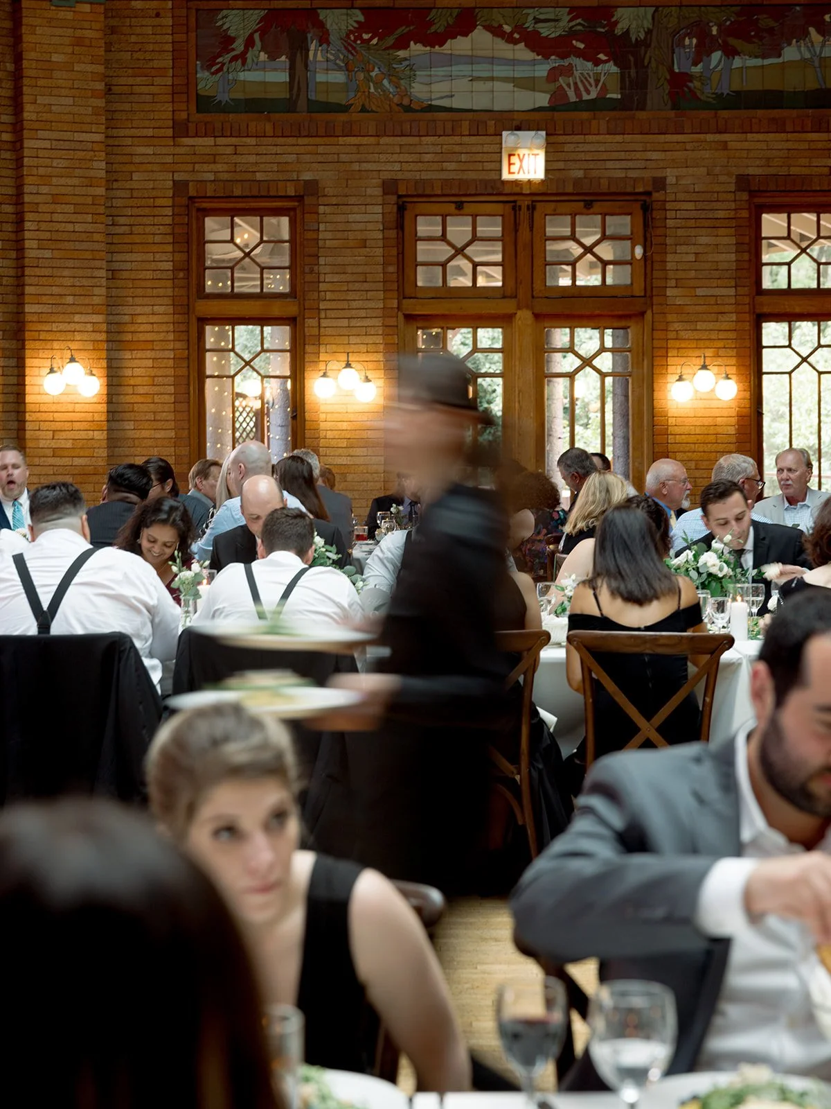Wide shot of Cafe Brauer's reception at full swing — guests dining, a server in warm motion blur, brick walls glowing.