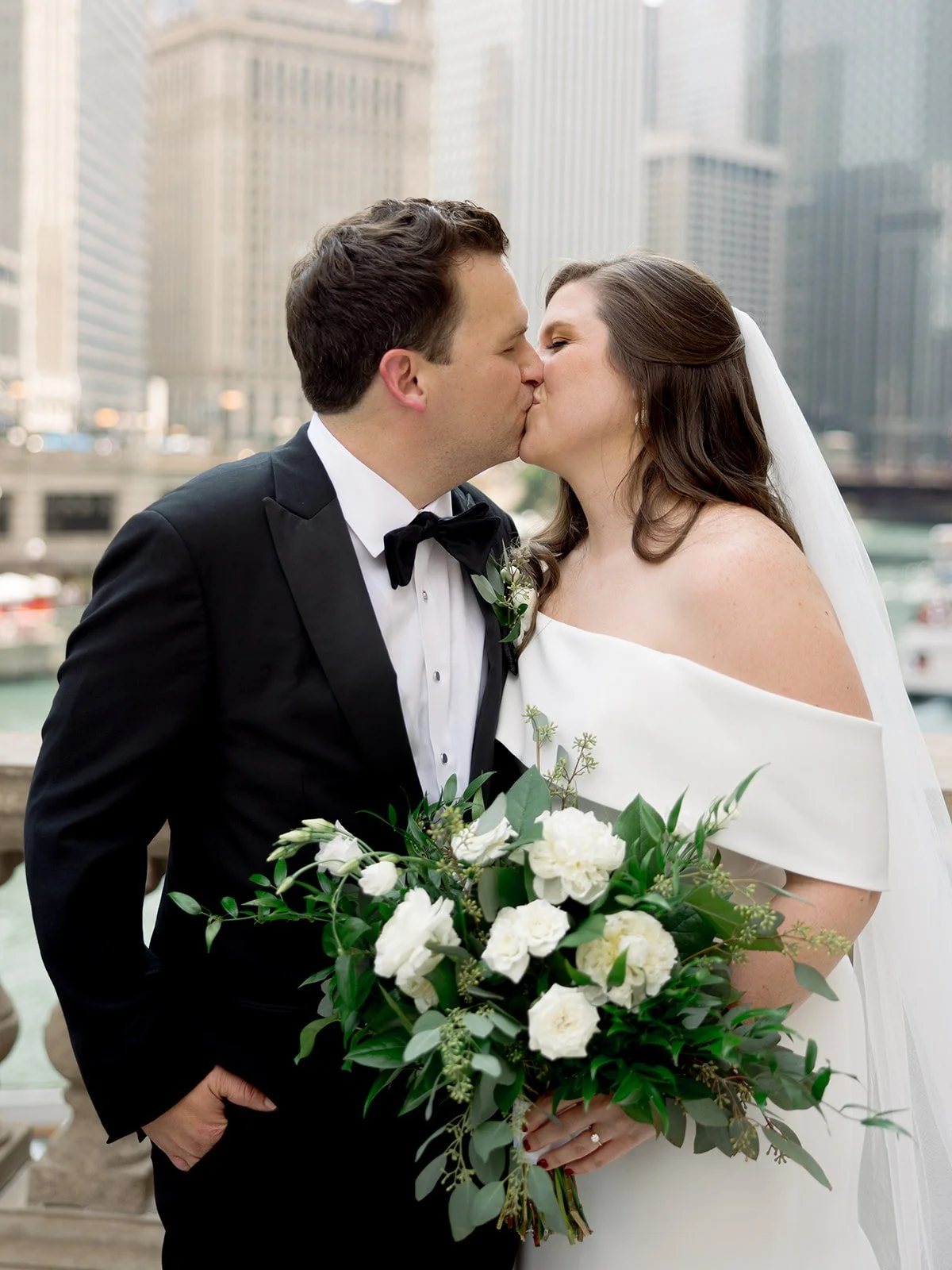 Bride and groom sharing a tender kiss on the Chicago Riverwalk, white bouquet between them, city skyline in the distance.