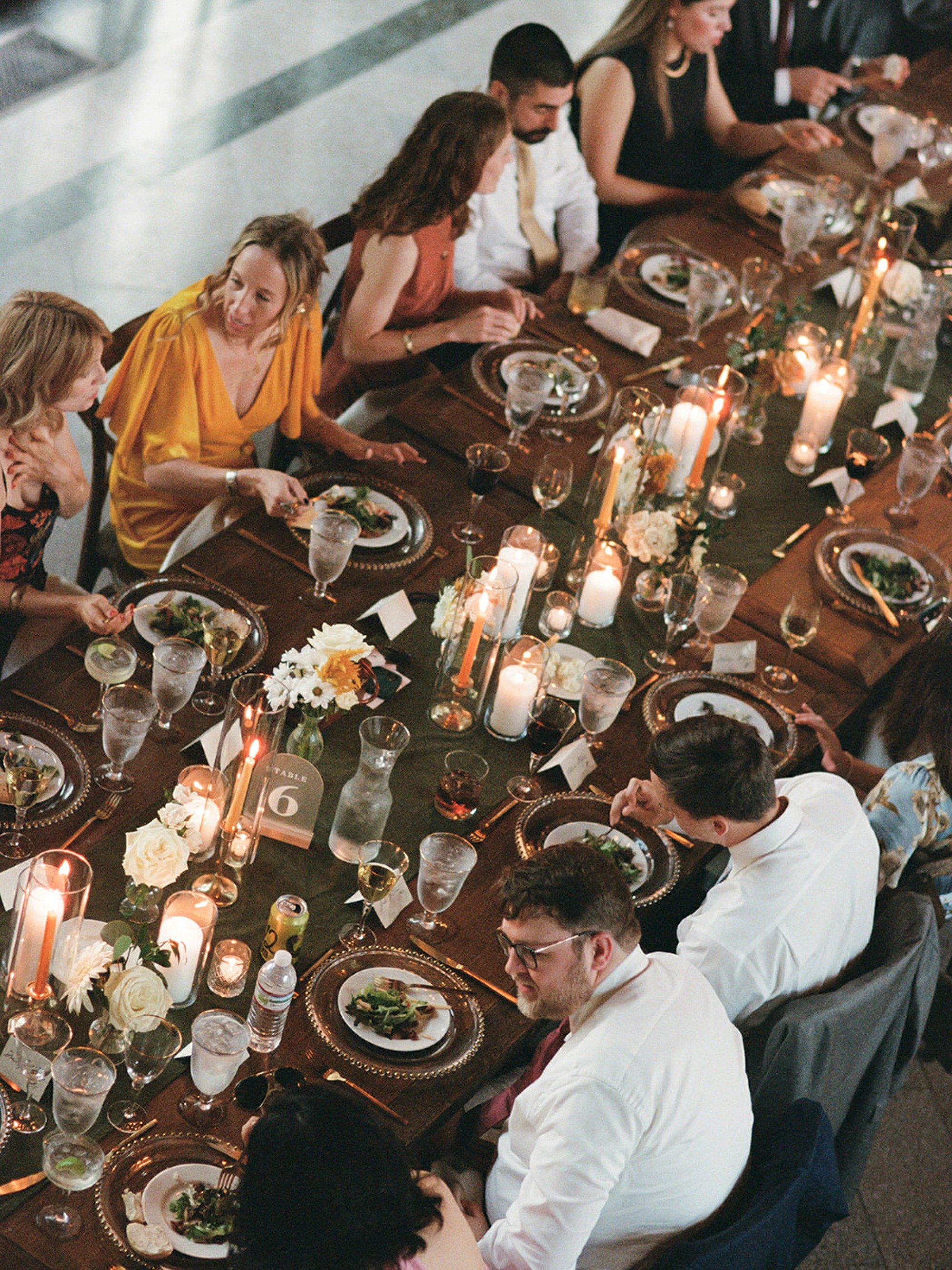 From above, guests gather around a farm table flickering with beeswax tapers and white florals — gold charger plates and crystal glassware completing the picture at this Columbus Park Refectory wedding