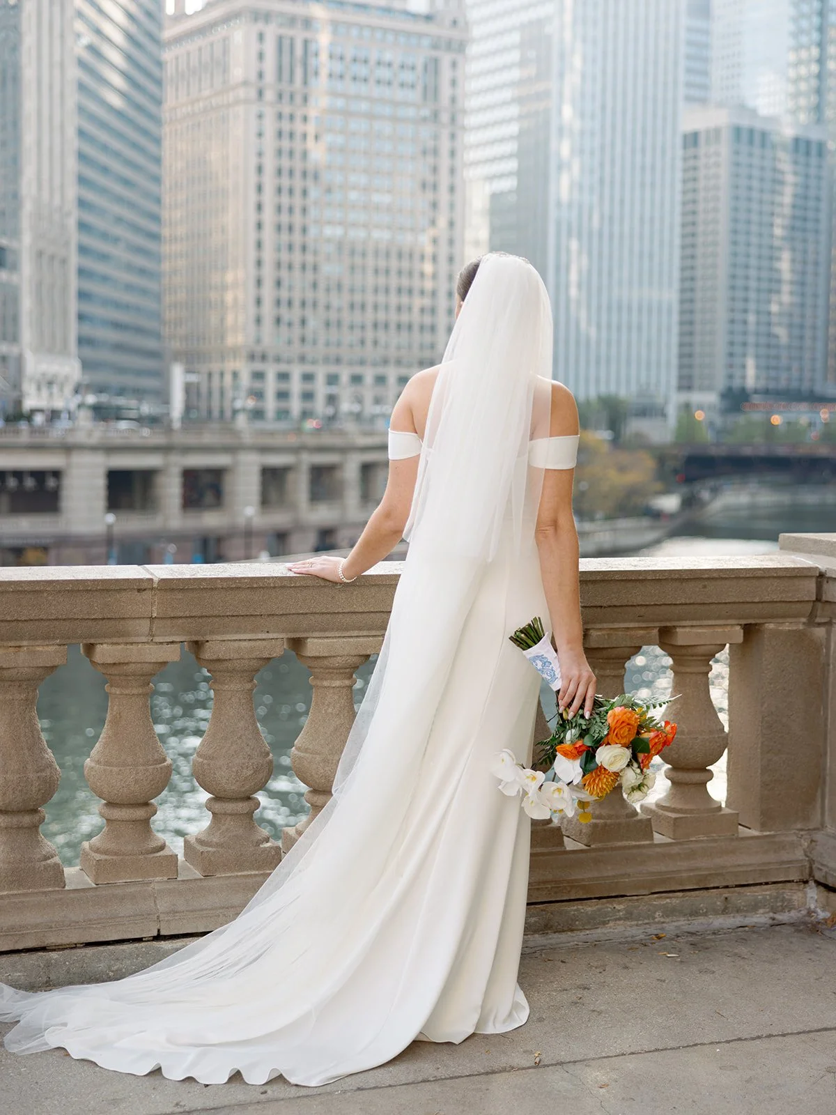 The bride gazes out over the Chicago River from the stone balustrade, cathedral veil cascading down her back as the downtown skyline fills every inch of the frame around her.