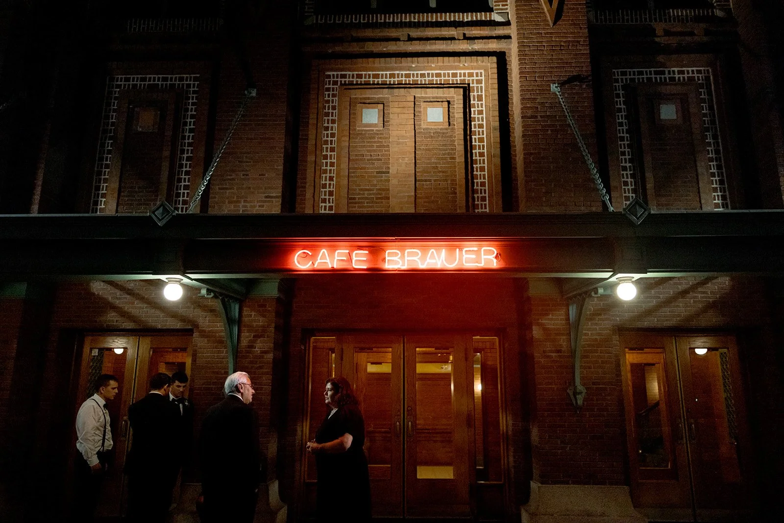 Cafe Brauer's exterior at night — neon sign casting warm red light over the brick facade, guests near the entrance.