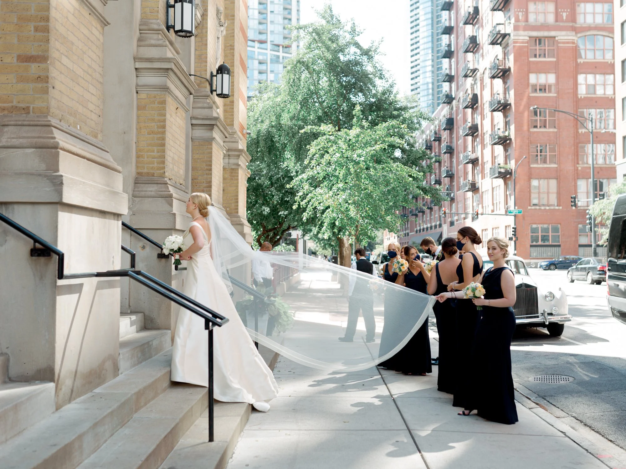 Bride stands poised at the top of stone church steps as her bridesmaids hold the full length of her cathedral veil along the Chicago sidewalk below.
