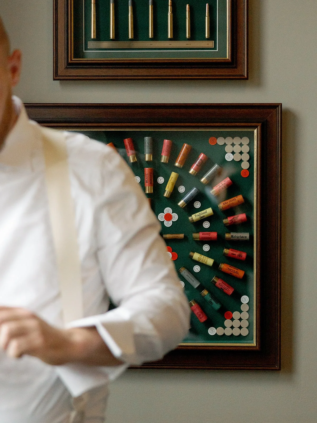 The groom adjusts his suspenders in front of a framed display of vintage shotgun shells — a character-filled getting-ready detail that tells you something real about who he is. Shot on film by a Chicago film wedding photographer.