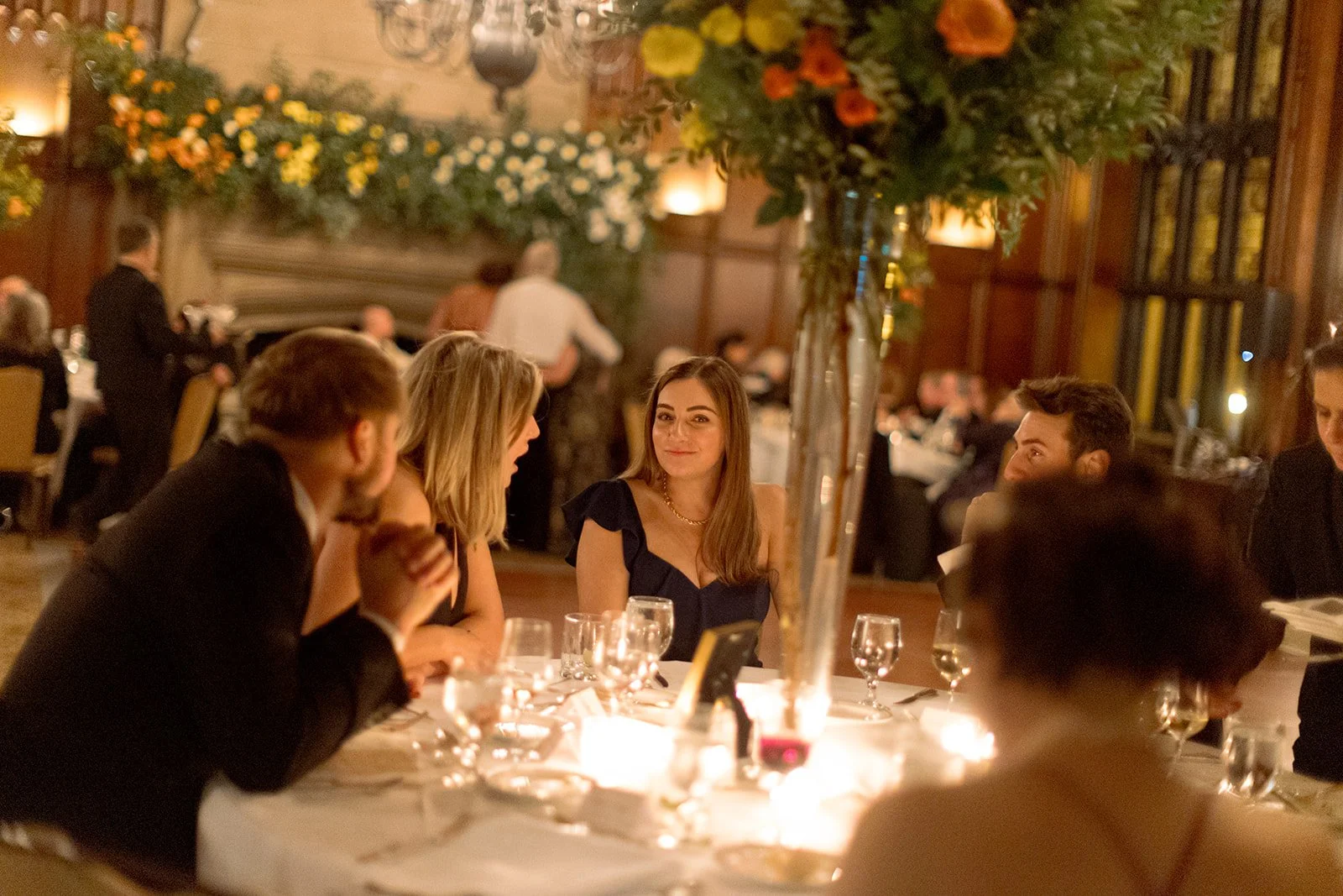 Guests deep in conversation at their candlelit tables, a tall floral centerpiece presiding over the scene, the warm ornate interior of University Club Chicago glowing all around.