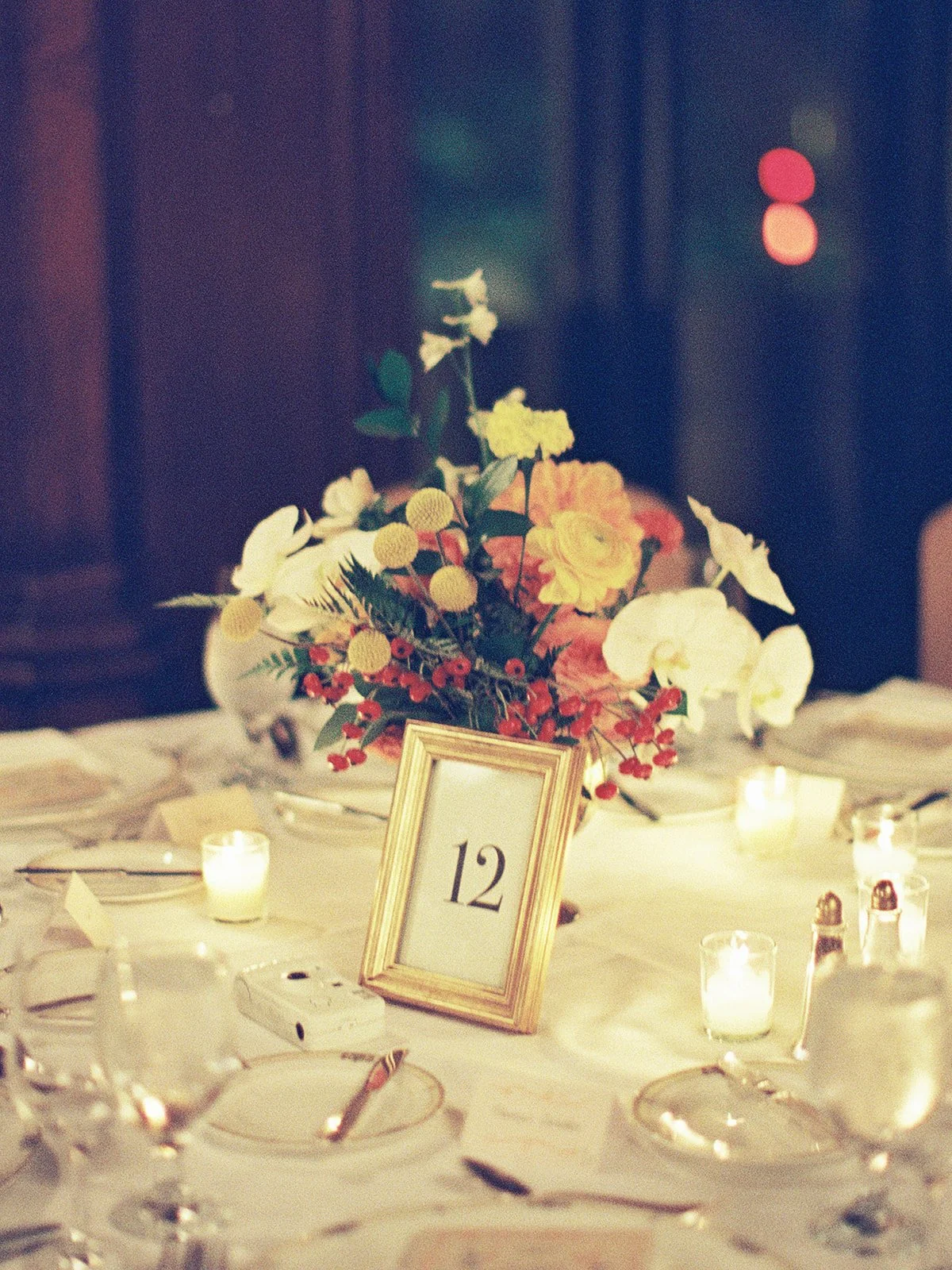 A low centerpiece of yellow, orange, and white blooms beside a gold-framed table number twelve, votives glowing around it on white linen. Warm, intimate reception table styling at University Club Chicago — by a Chicago film wedding photographer.