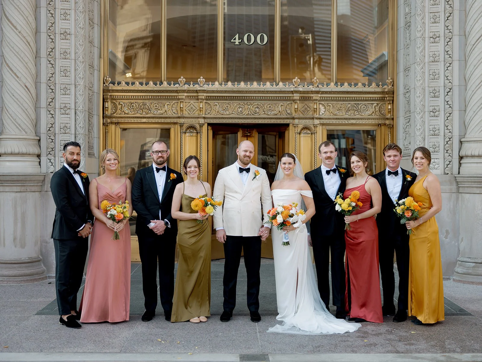 The full wedding party posed in front of Chicago's gold-gilded 400 South Michigan entrance — earth-tone bridesmaids and black-tuxedoed groomsmen flanking the couple in a crisp, architectural group portrait.