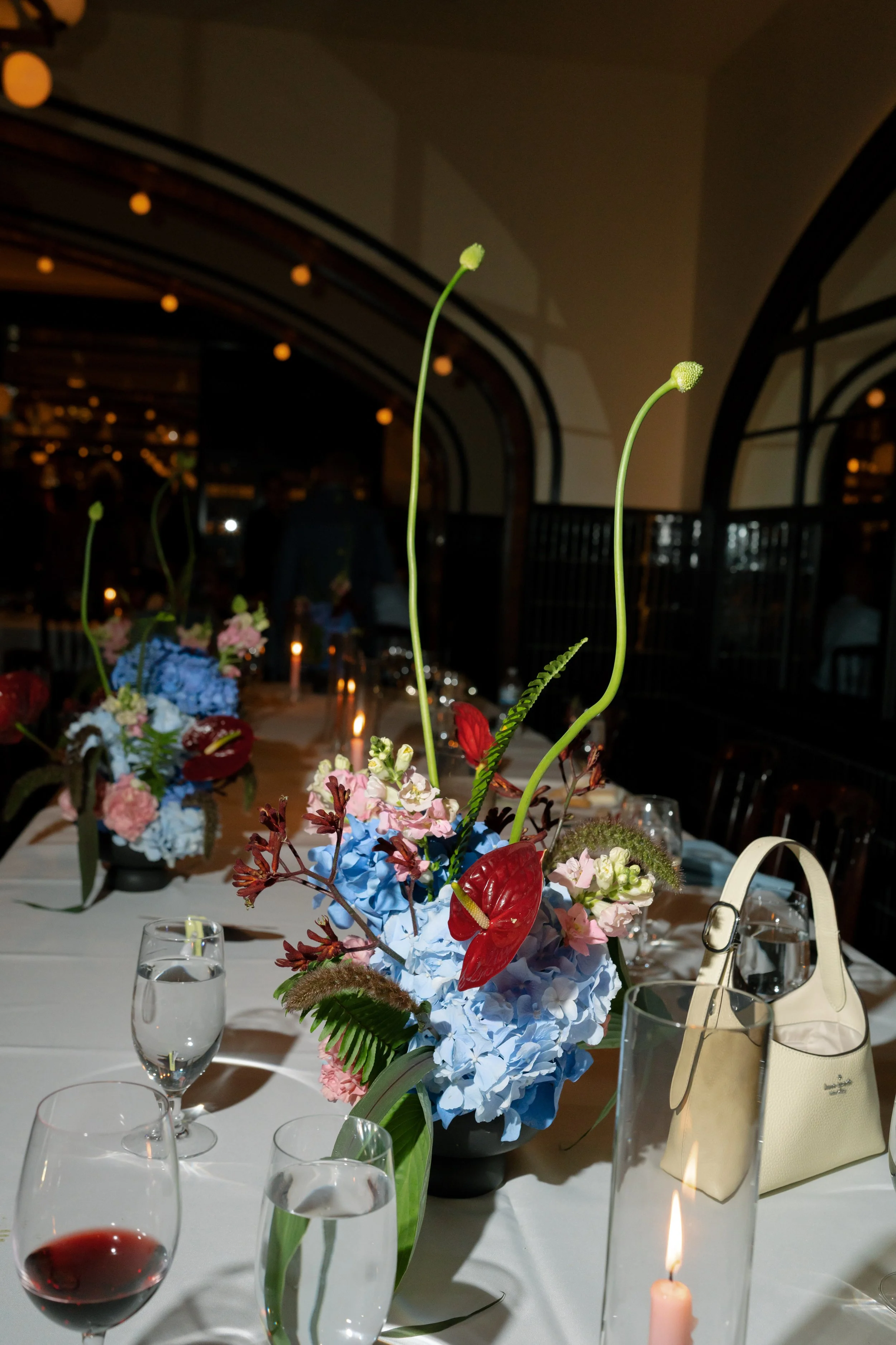 Blue hydrangea and red anthurium centerpiece glows on a candlelit reception table beneath arched windows