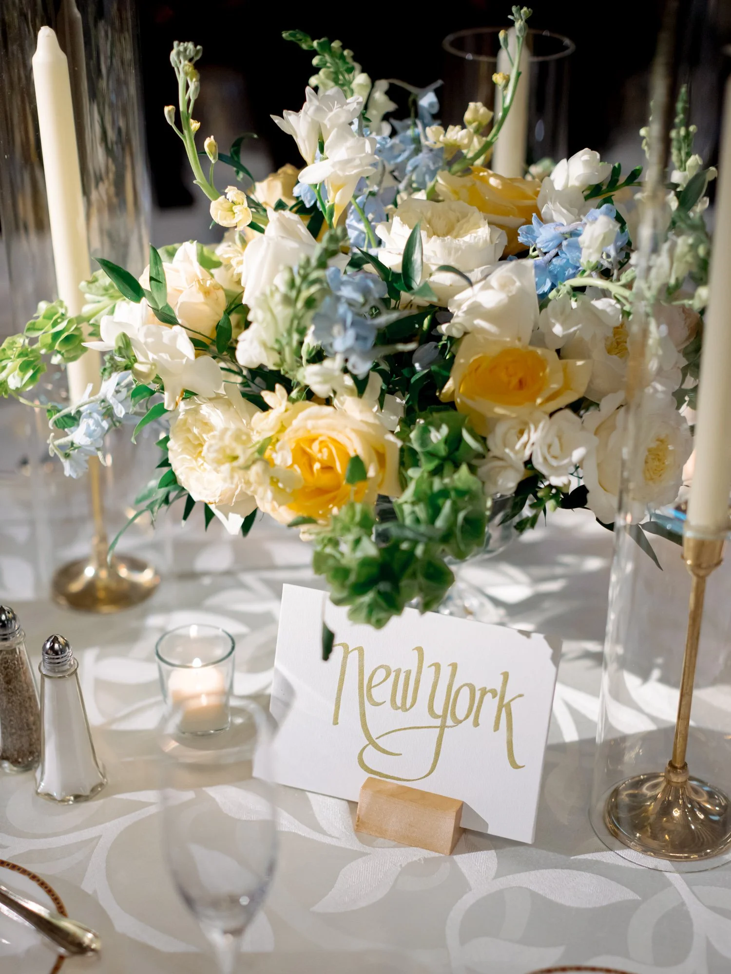 A table card reading ‘New York’ in gold script rests beside yellow roses and blue hydrangea — city-named tables, a detail both personal and polished.
