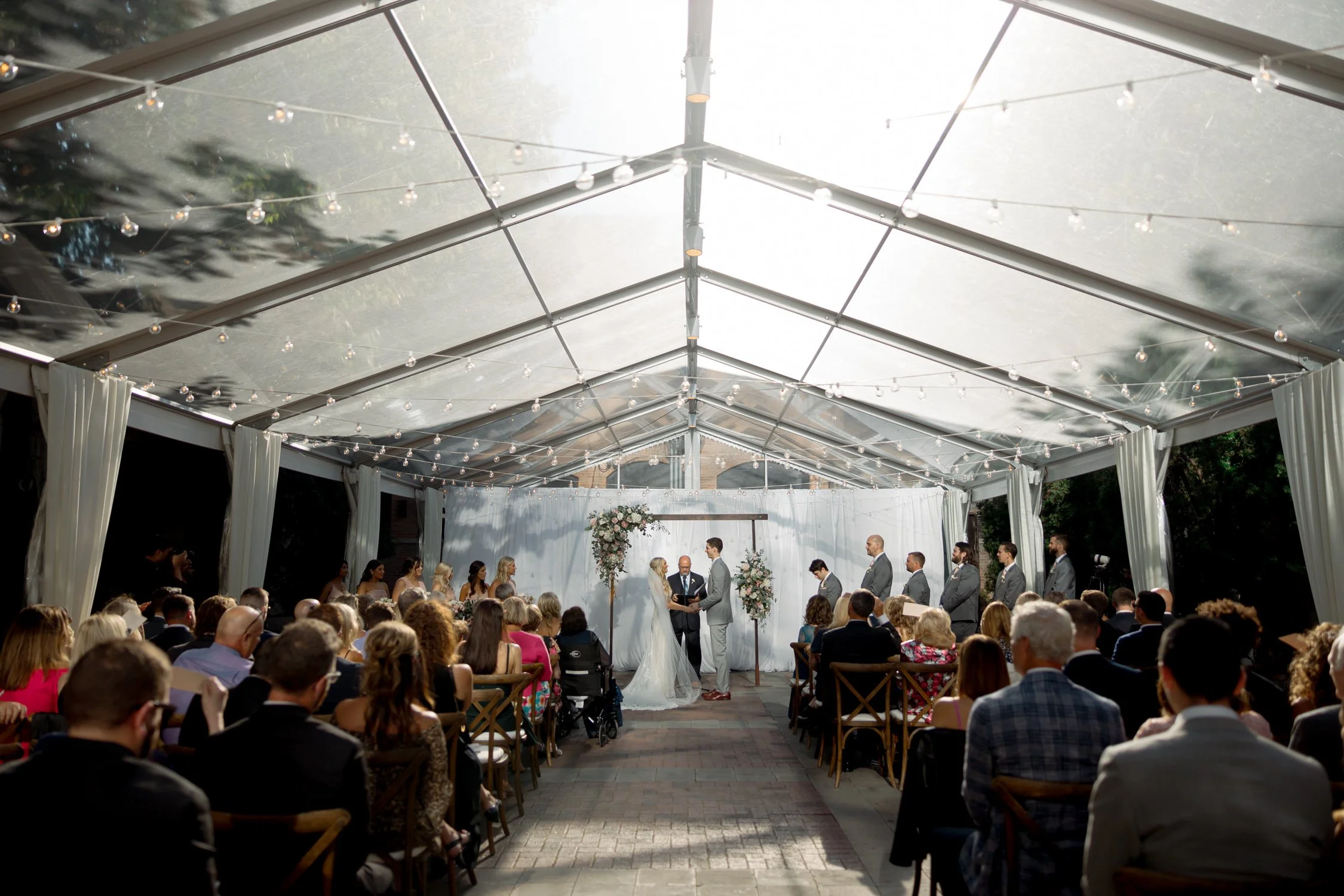 A breathtaking wide shot of the full wedding ceremony under the glass-roofed tent at the Chicago Illuminating Company, string lights glowing above the crowd.