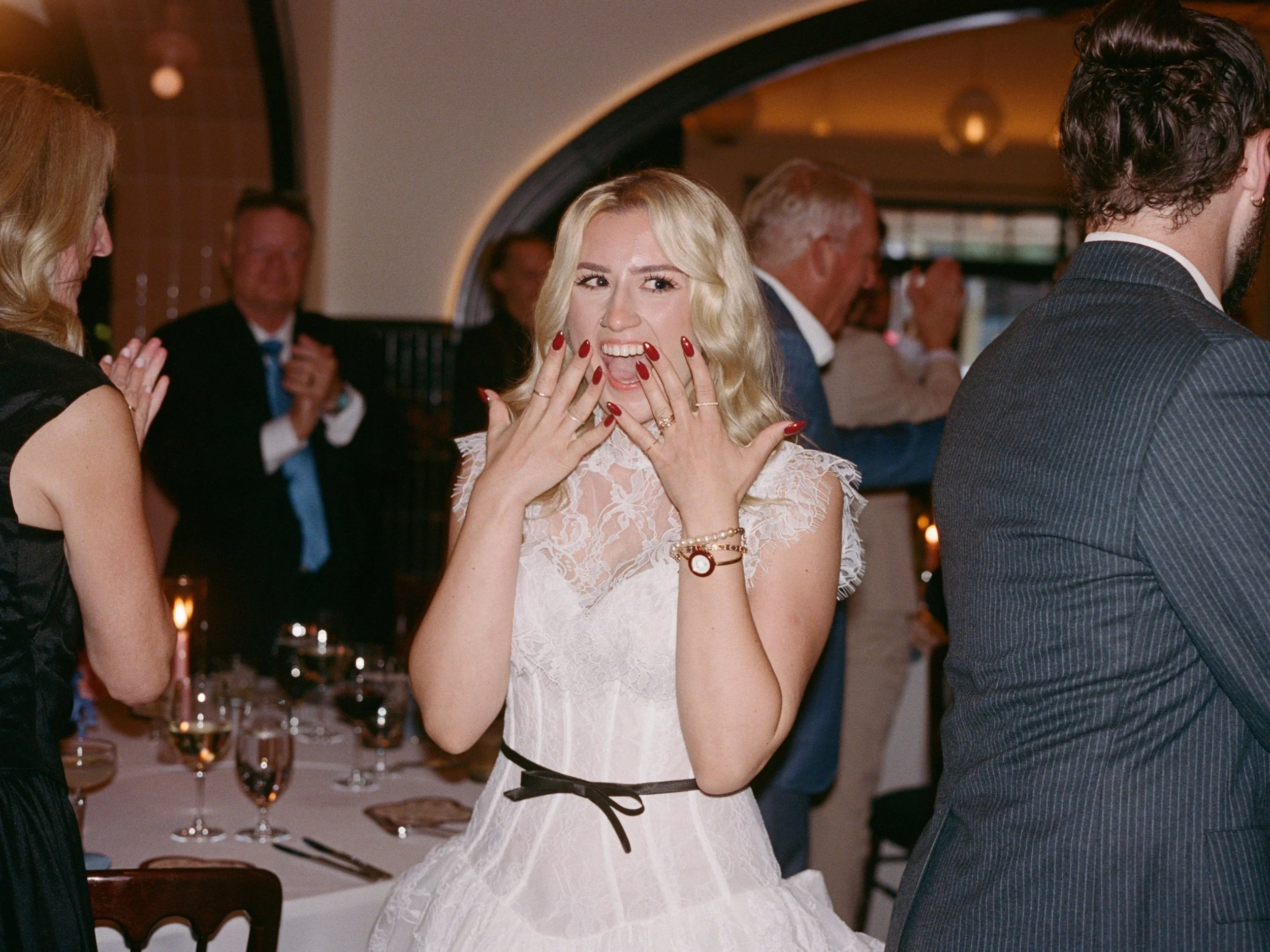 Bride laughs with hands raised at the reception table surrounded by guests