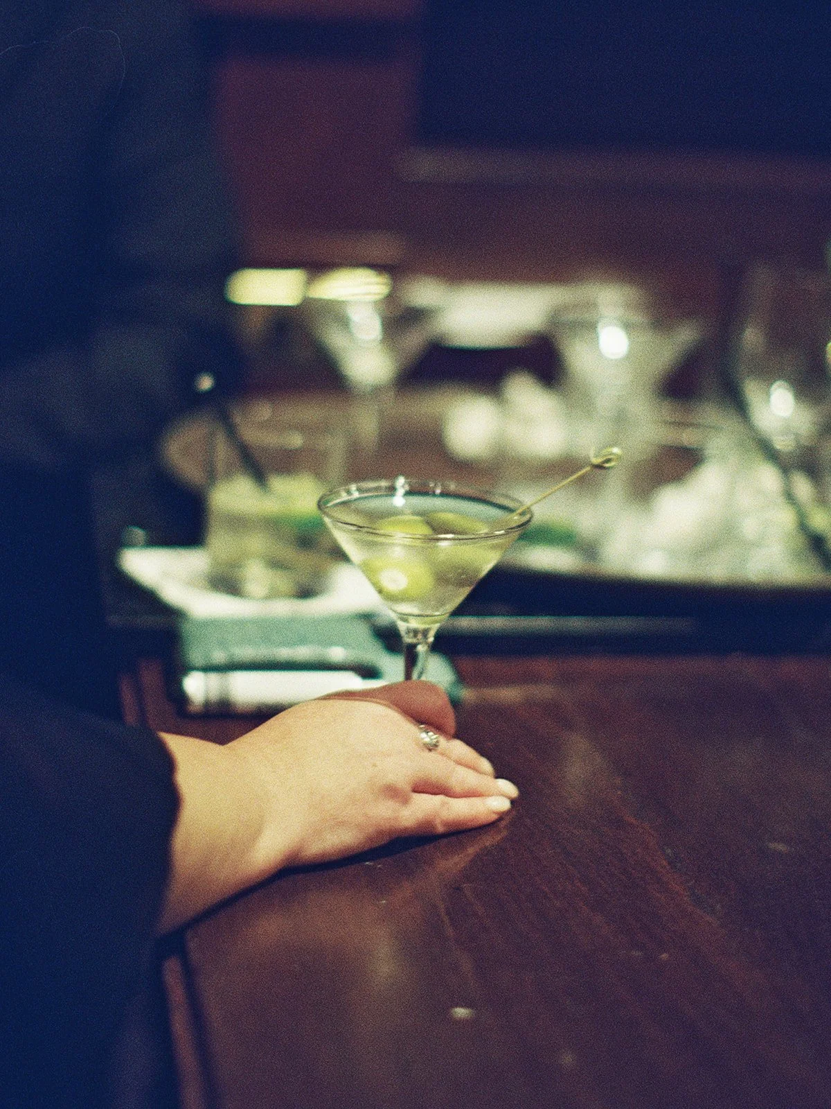 A hand rests on the dark bar top beside a classic dirty martini, three olives skewered and waiting — moody, intimate, and unmistakably film. The University Club bar at cocktail hour, by a Chicago film wedding photographer.