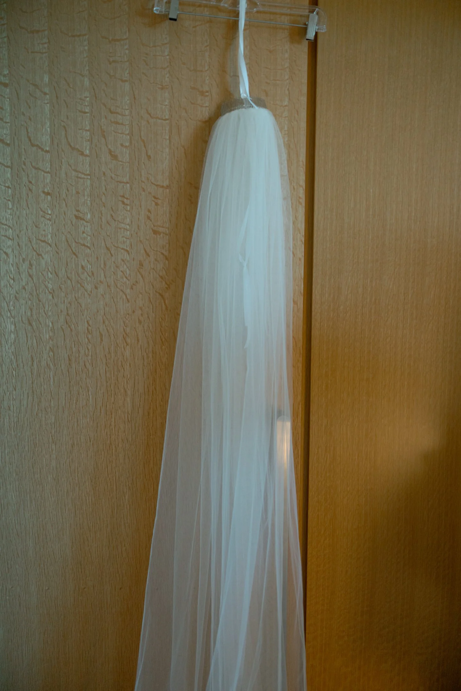 The bride's long cathedral veil hangs softly from a hotel closet rod, its sheer tulle cascading in delicate folds before the wedding day begins.