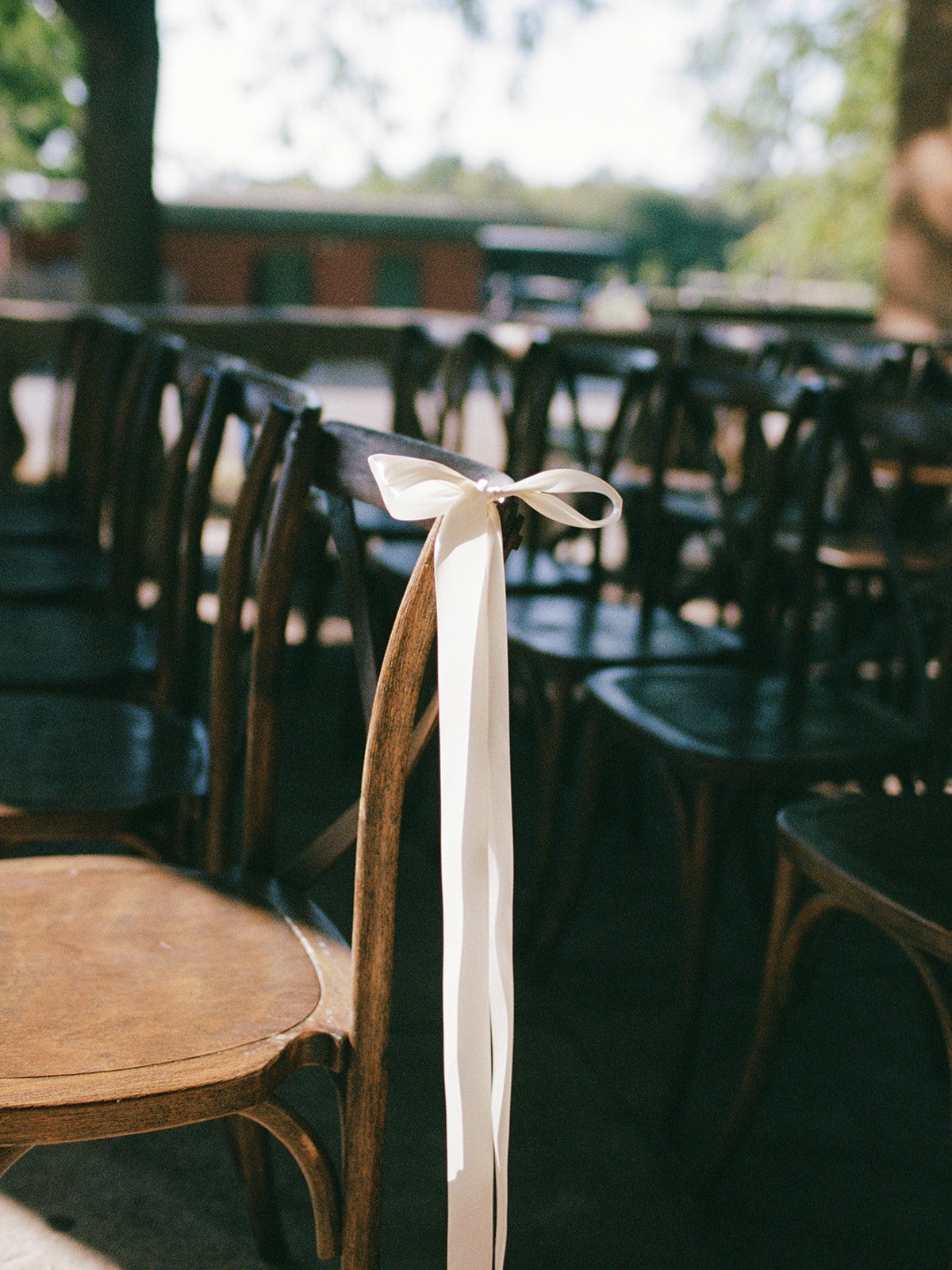 A simple, elegant detail — a long white silk ribbon tied in a bow on a wooden cross-back ceremony chair, rows of seats awaiting guests in soft dappled sunlight at Columbus Park Refectory. Chicago film wedding photographer.